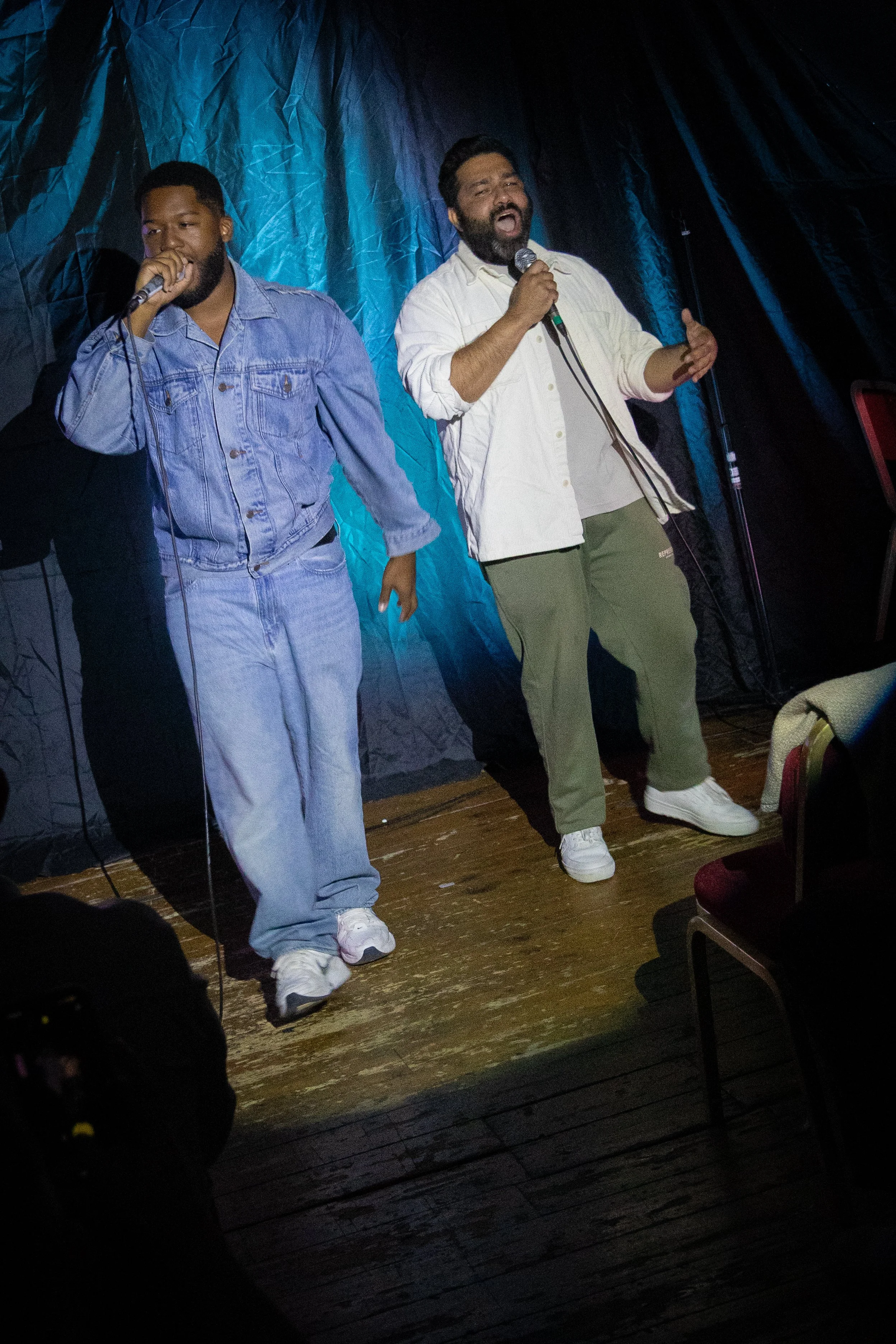 Two men singing on a stage, one with a microphone and wearing a light-colored shirt and green pants, the other in a denim jacket and jeans, with a dark background and some chairs visible.