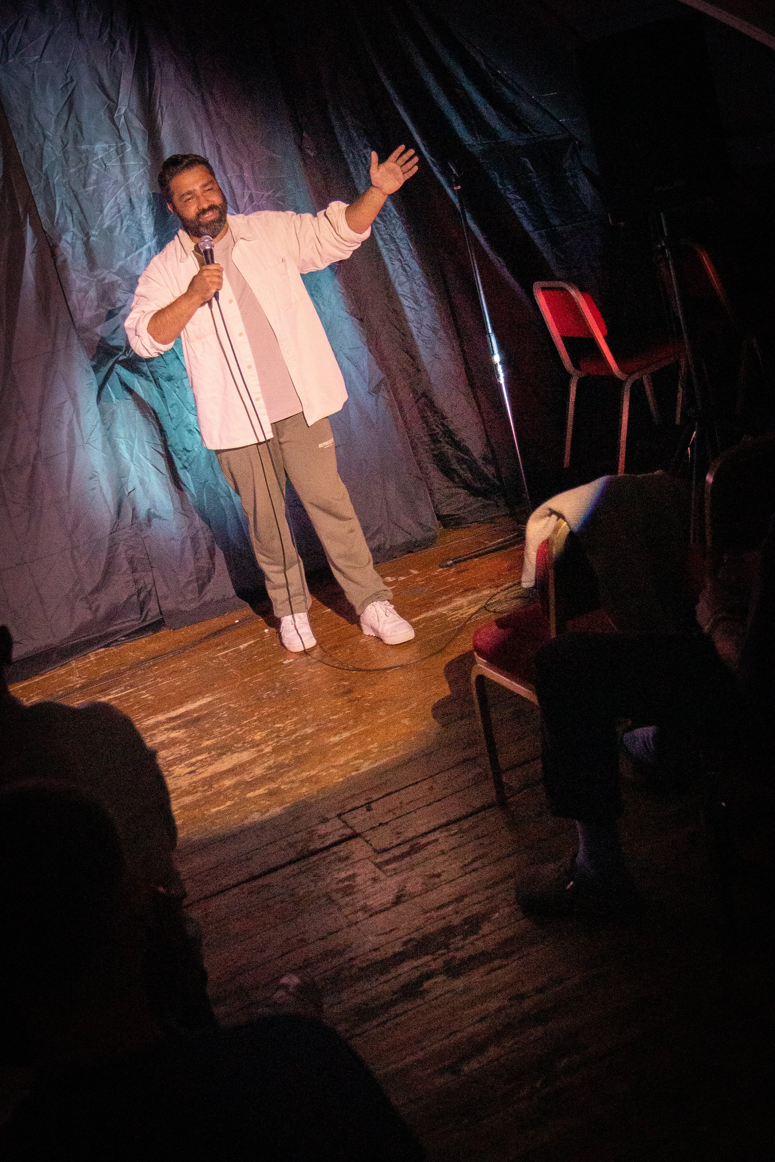 A man with a beard, wearing a light pink shirt and beige pants, is standing on a small stage with a dark backdrop, holding a microphone and gesturing with his left hand. The stage has a wooden floor and black curtains, with an empty red chair nearby.
