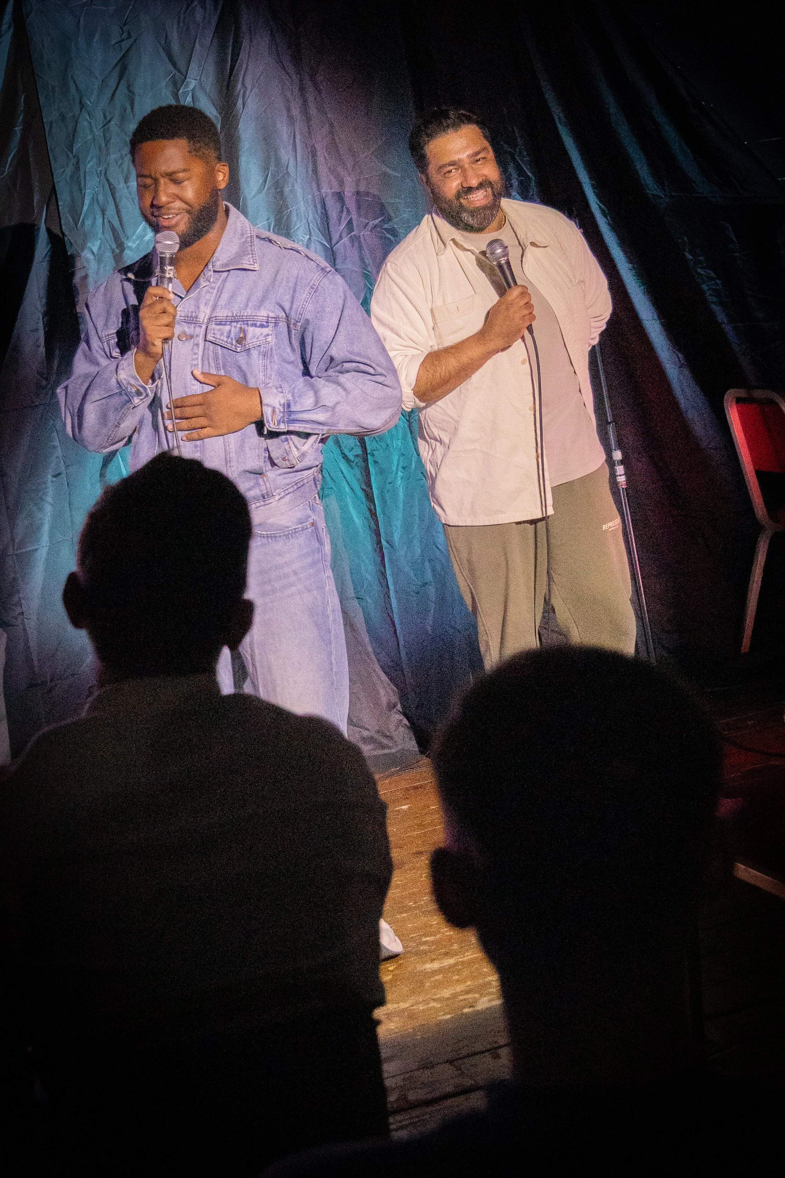 Two men performing stand-up comedy on a dimly lit stage, holding microphones, with an audience sitting in front of them.