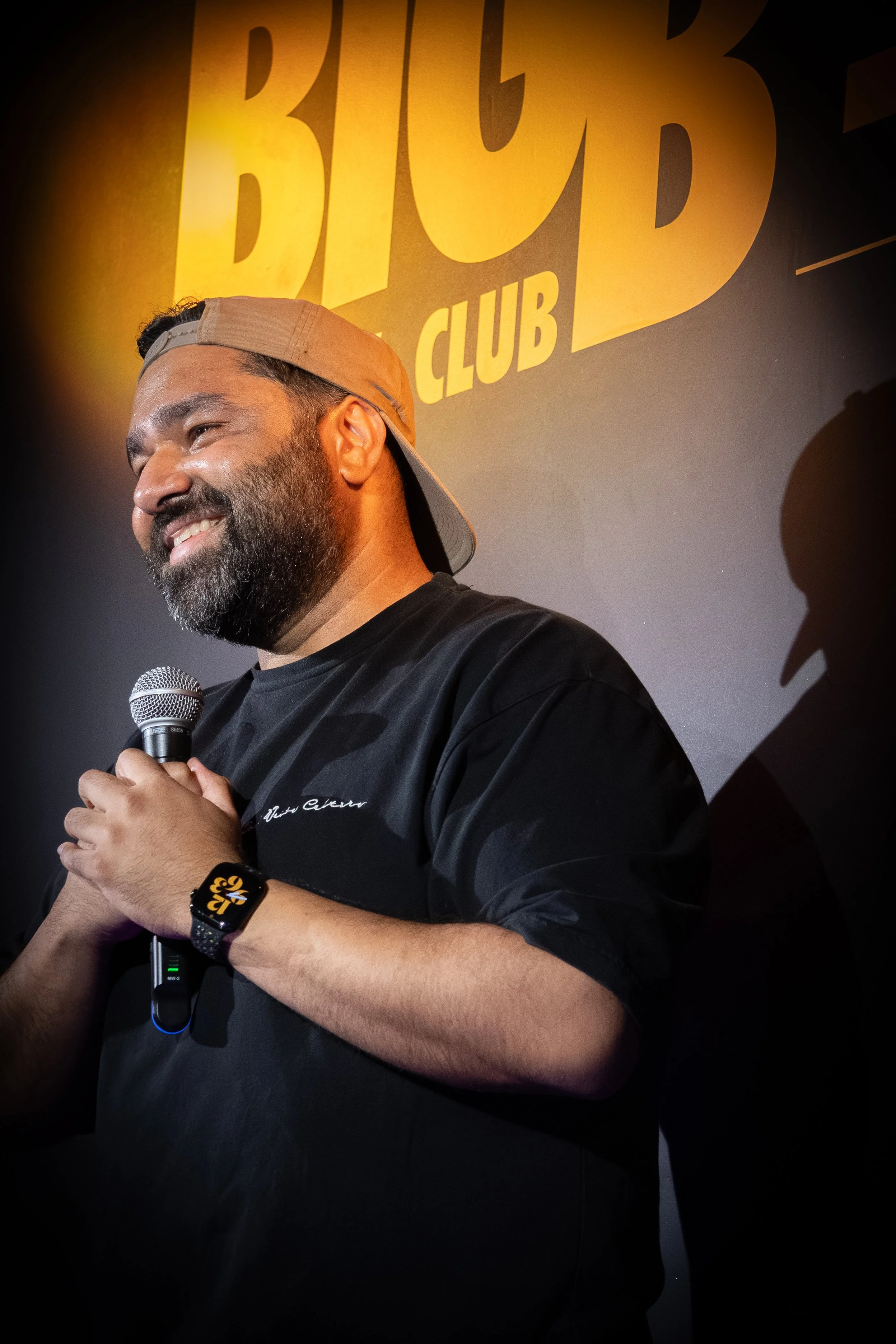 A man with a beard wearing a backwards tan cap and a black T-shirt, smiling and holding a microphone, standing in front of a yellow-lit sign that reads 'Big Belly Comedy Club"' in bold letters.