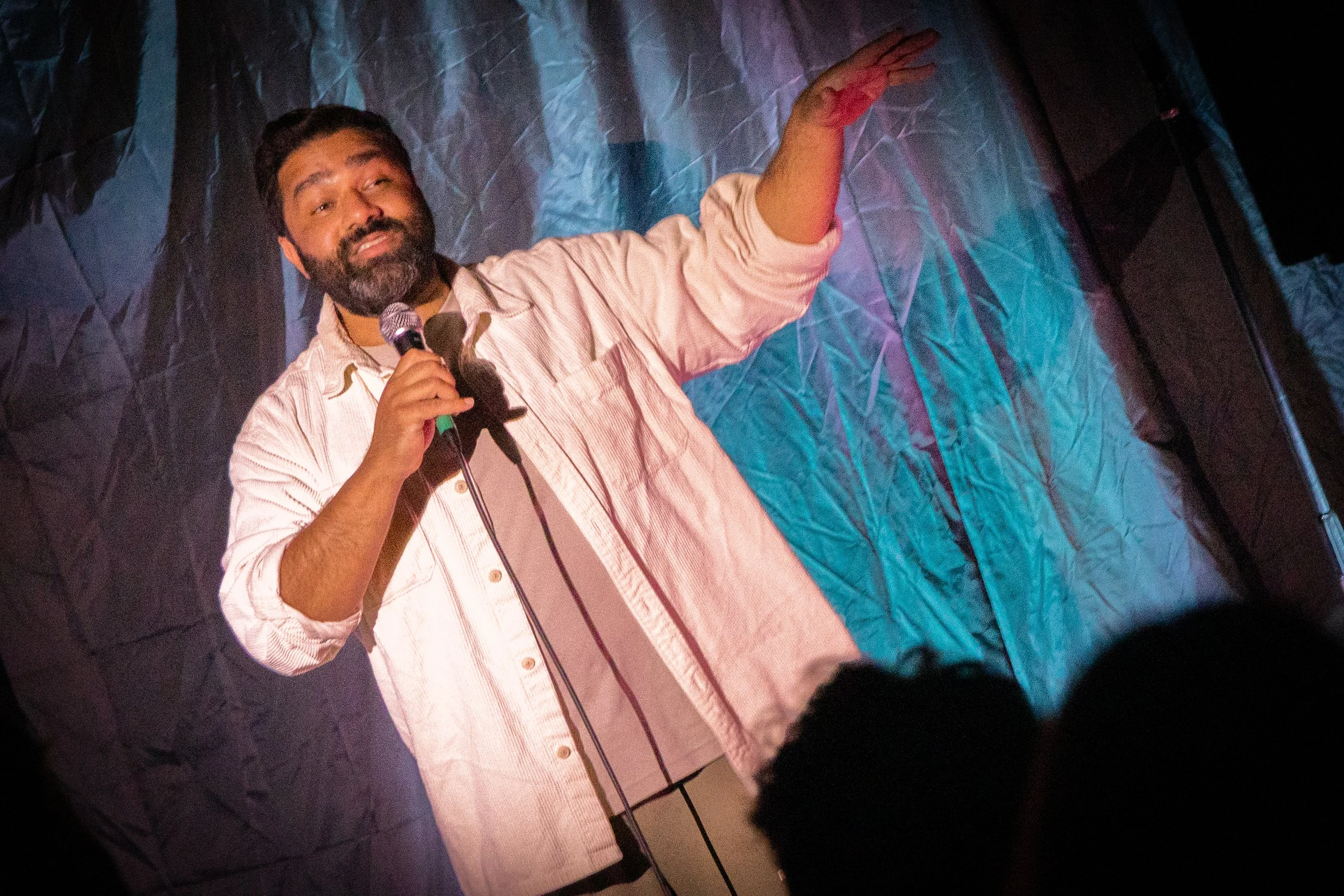 A man with a beard and dark hair holding a microphone, speaking or singing on stage with a colorful background and dramatic lighting.