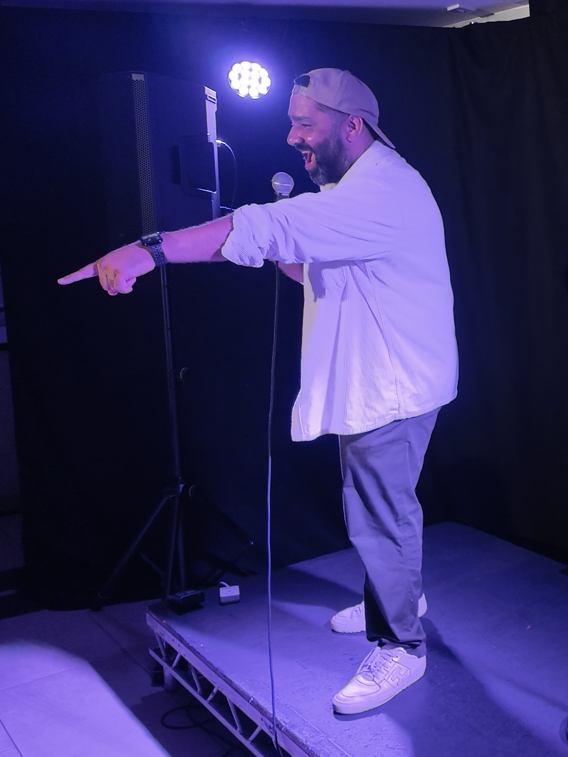 A man with a beard wearing a backwards baseball cap, white jacket, and gray pants is standing on a small platform, pointing and singing or speaking into a microphone with purple stage lighting in the background.