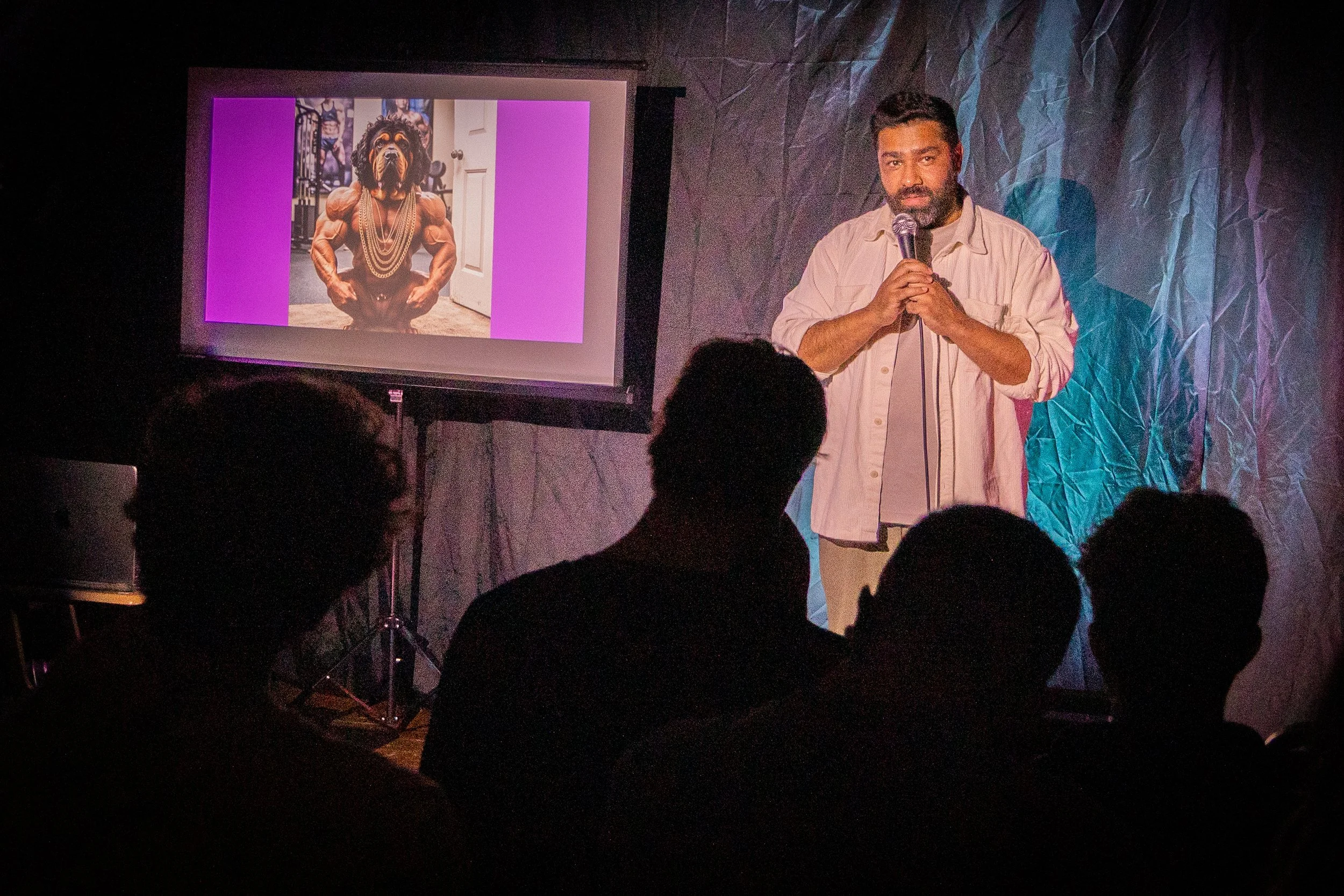 A man with a beard holding a microphone, standing in front of a dark background with crumpled paper texture, and a projection screen displaying an animated muscular dog with a human body at a presentation or event.