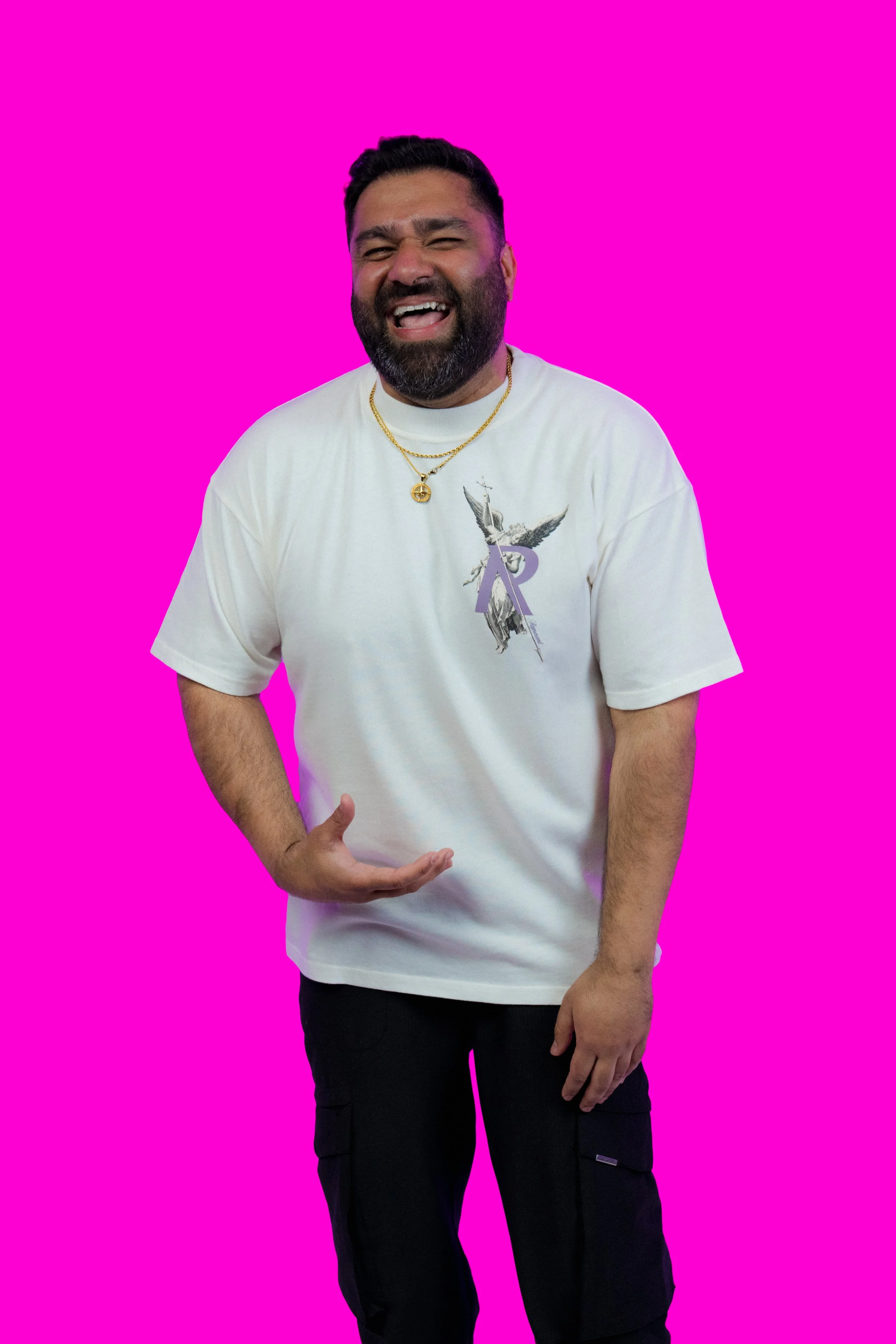 A man with dark hair and a beard laughing, wearing a white T-shirt with a graphic print and gold necklaces, standing against a bright pink background.