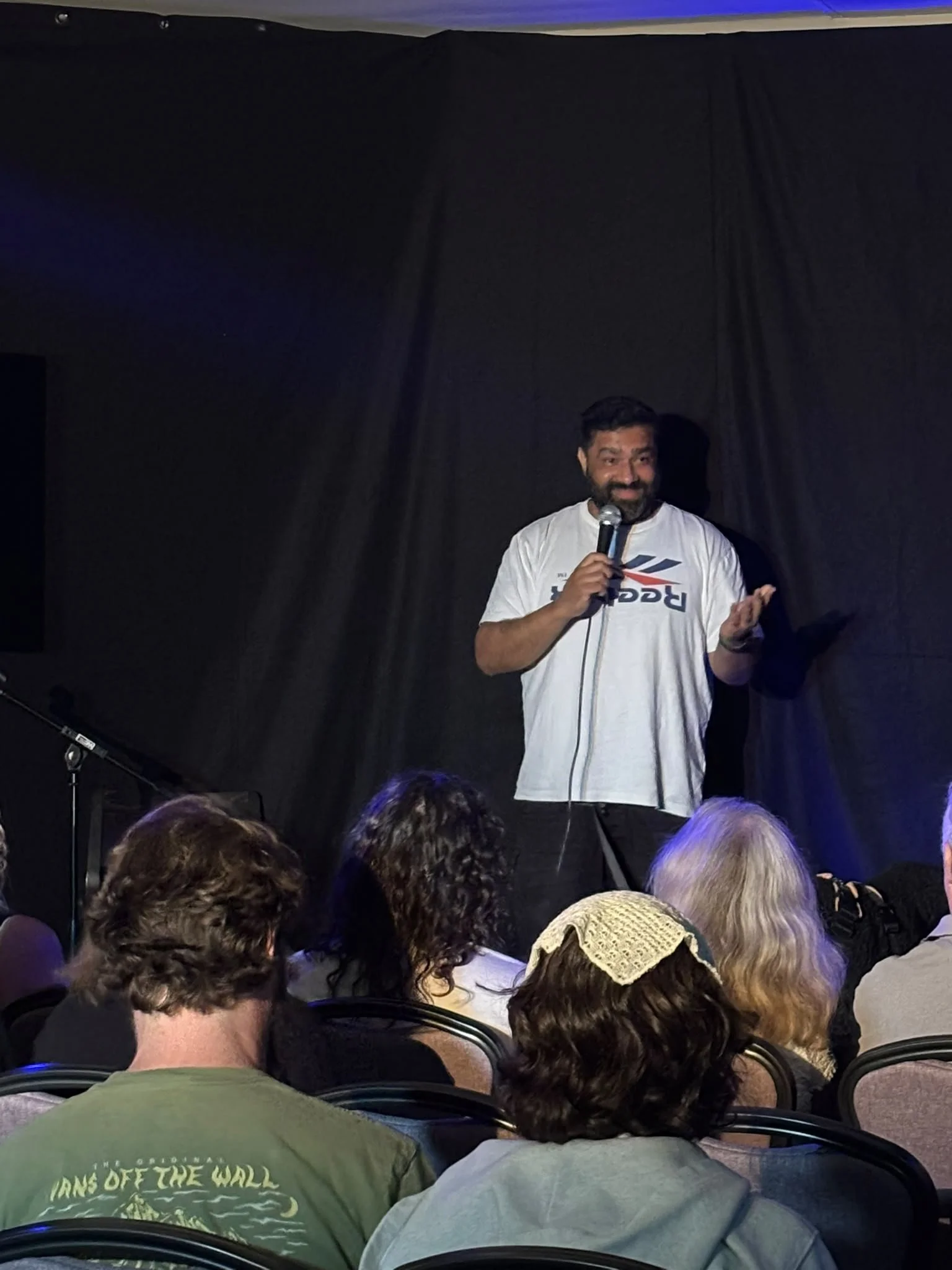 A man with a beard and mustache speaking into a microphone on stage in front of a dark curtain, audience members seated in front of him.