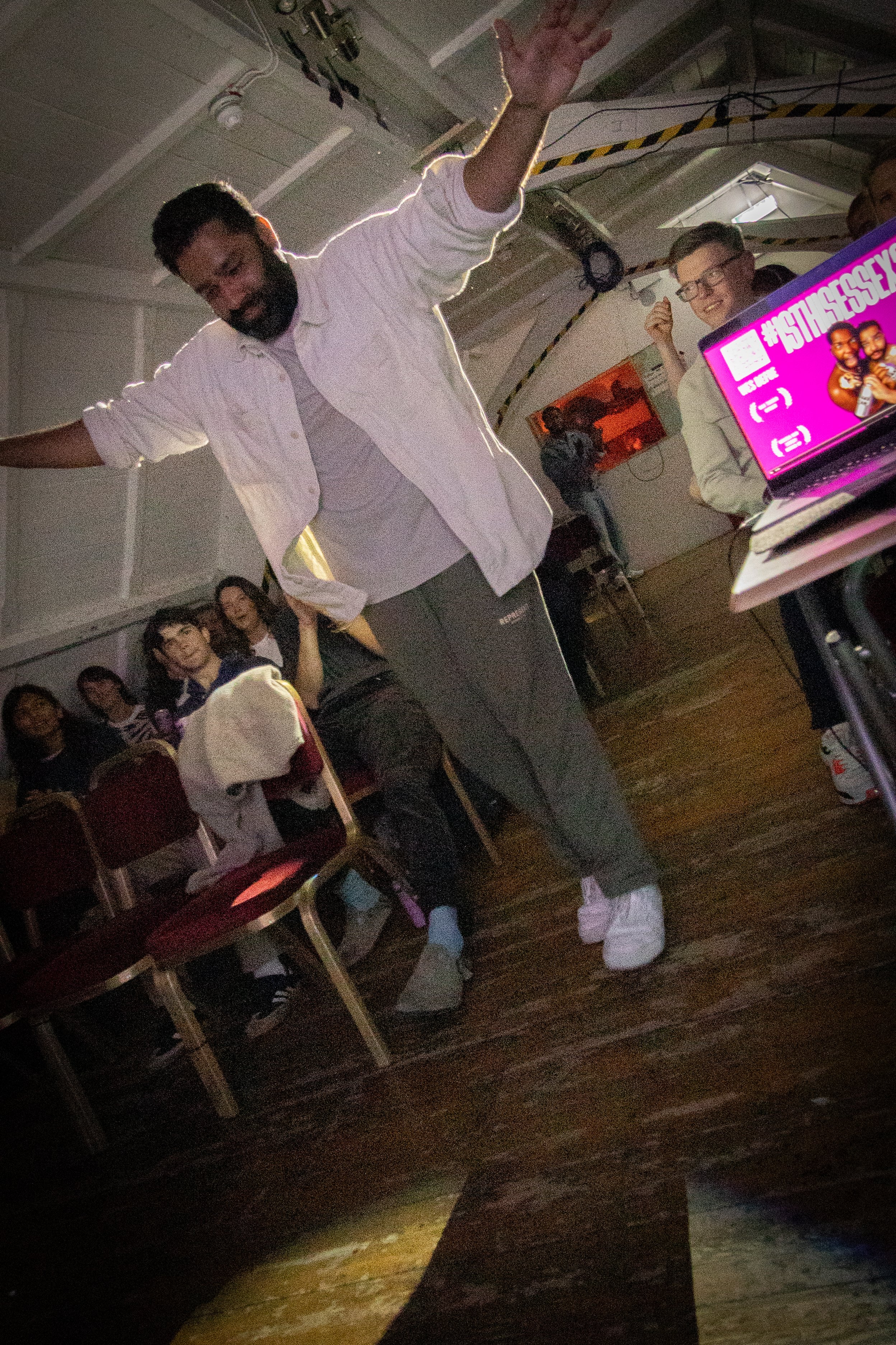A man with a beard and dreadlocks wearing a white shirt and grey pants is dancing with his arms raised in a dimly lit room with a wooden floor. There are several people in the background, including a woman wearing glasses and a man with a backpack. T