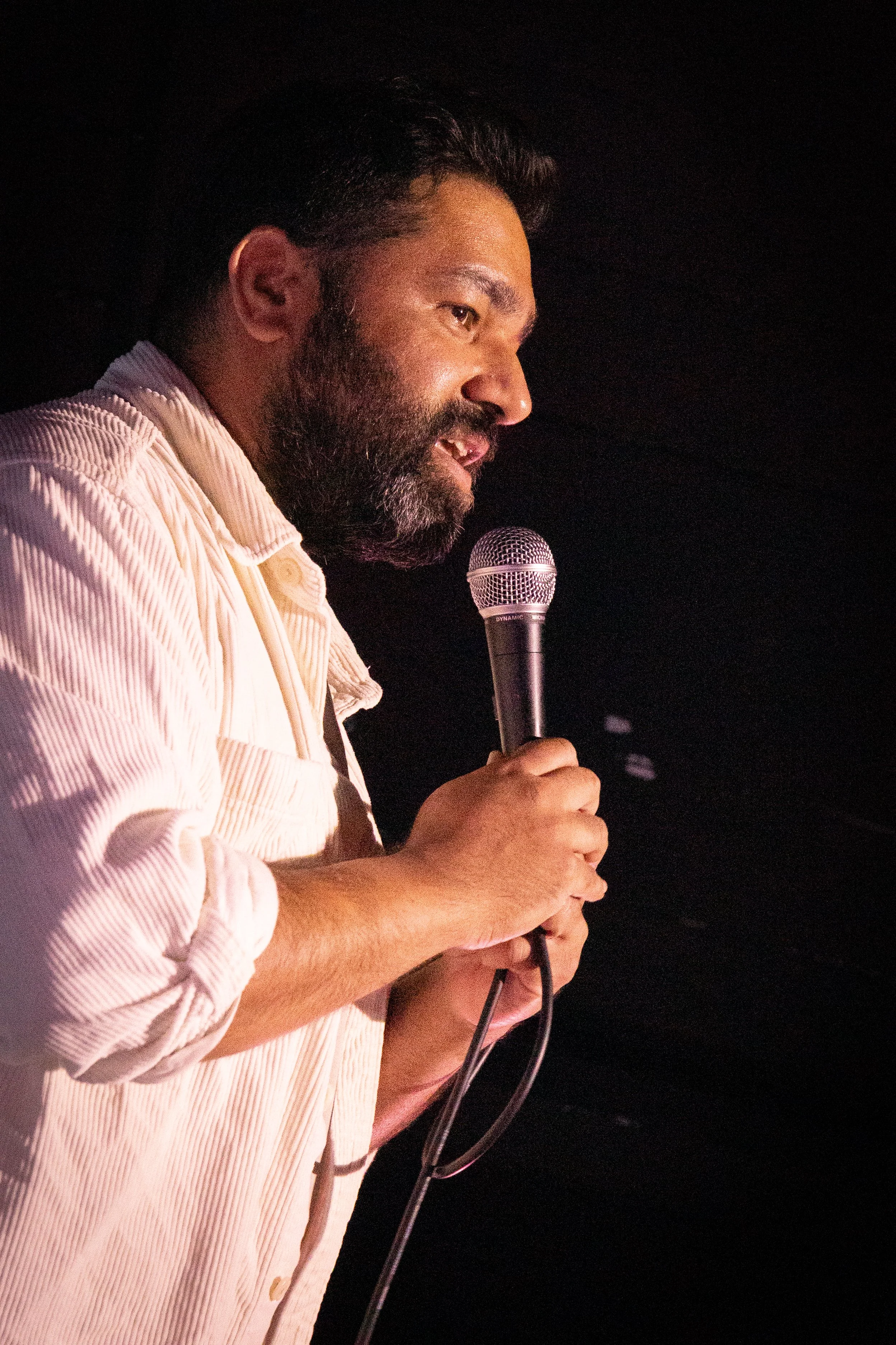 A man holding a microphone and speaking or performing on stage, wearing a light-colored, striped, button-up shirt.