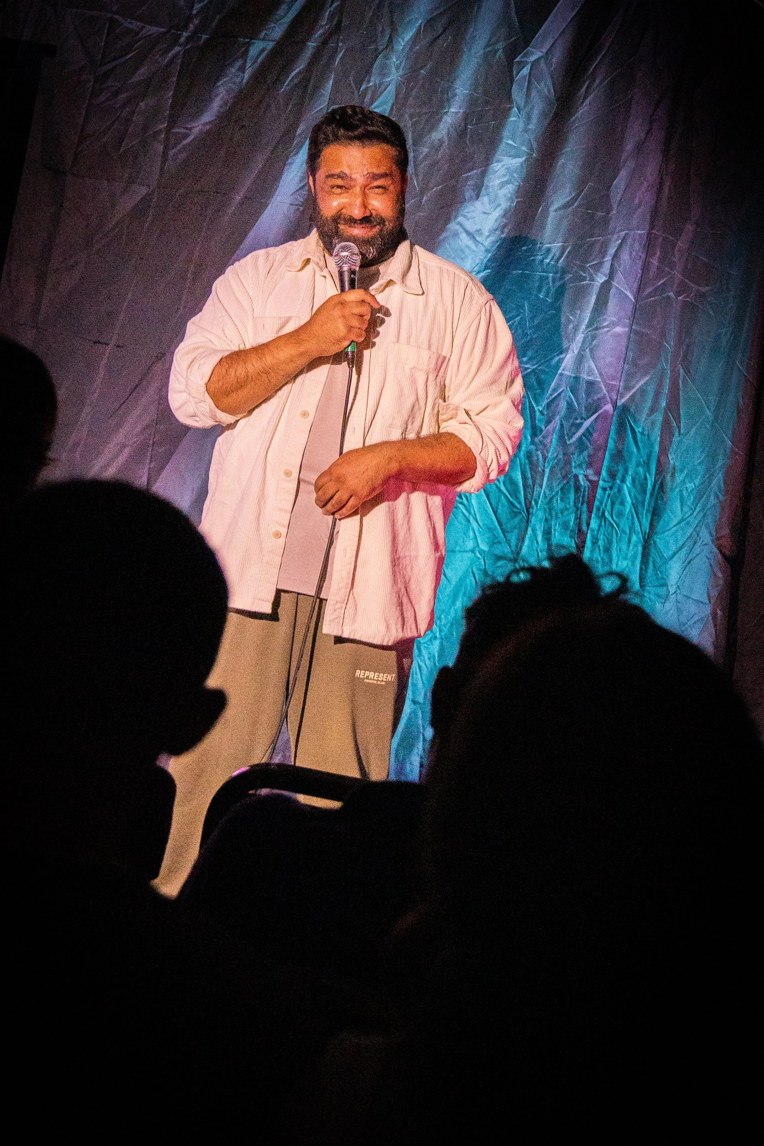 A man with a beard holding a microphone on stage, smiling, with a crumpled blue curtain backdrop, audience silhouettes in the foreground.