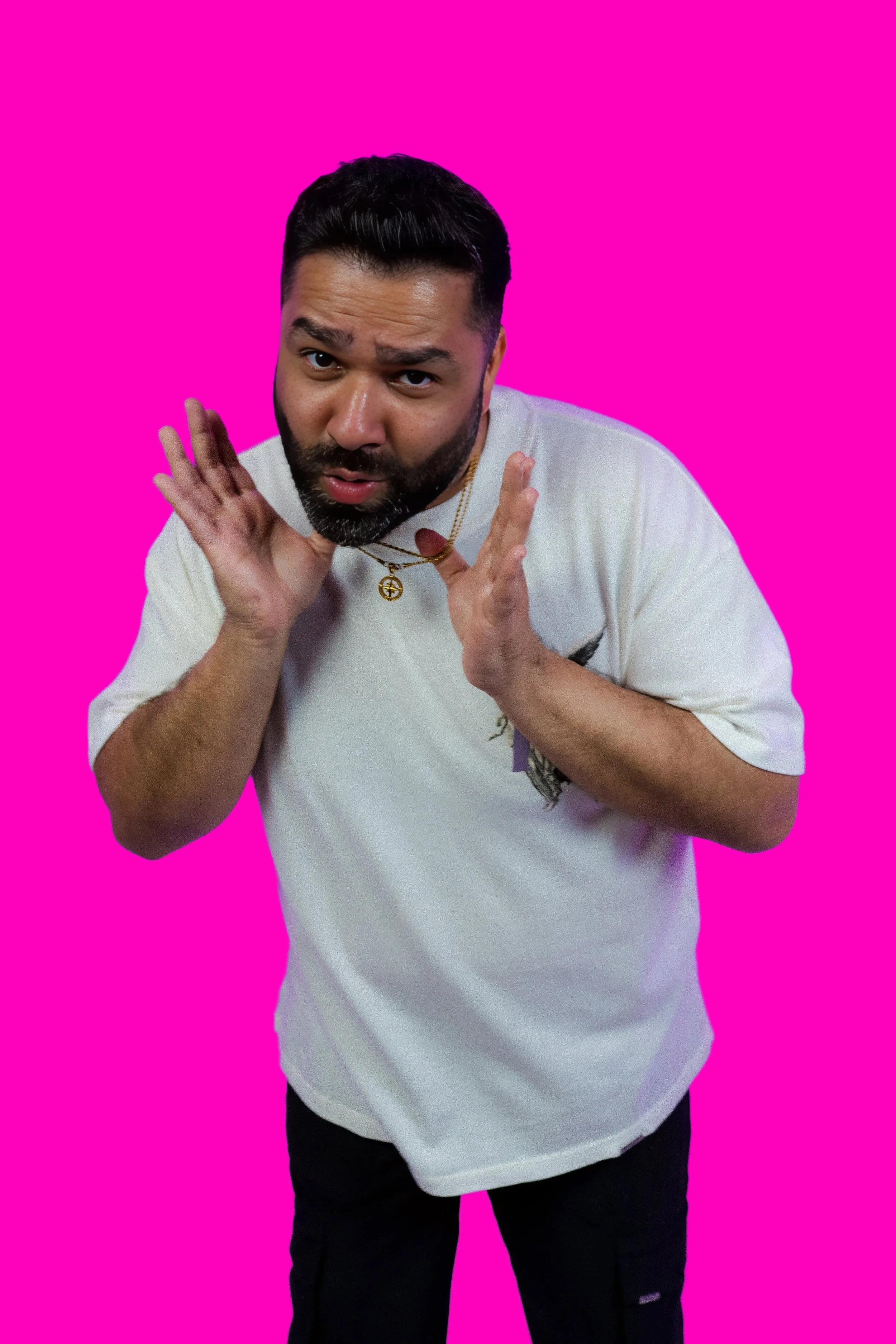 A man with dark hair and a beard is gesturing with his hands near his face, wearing a white t-shirt and gold jewelry, against a bright pink background.