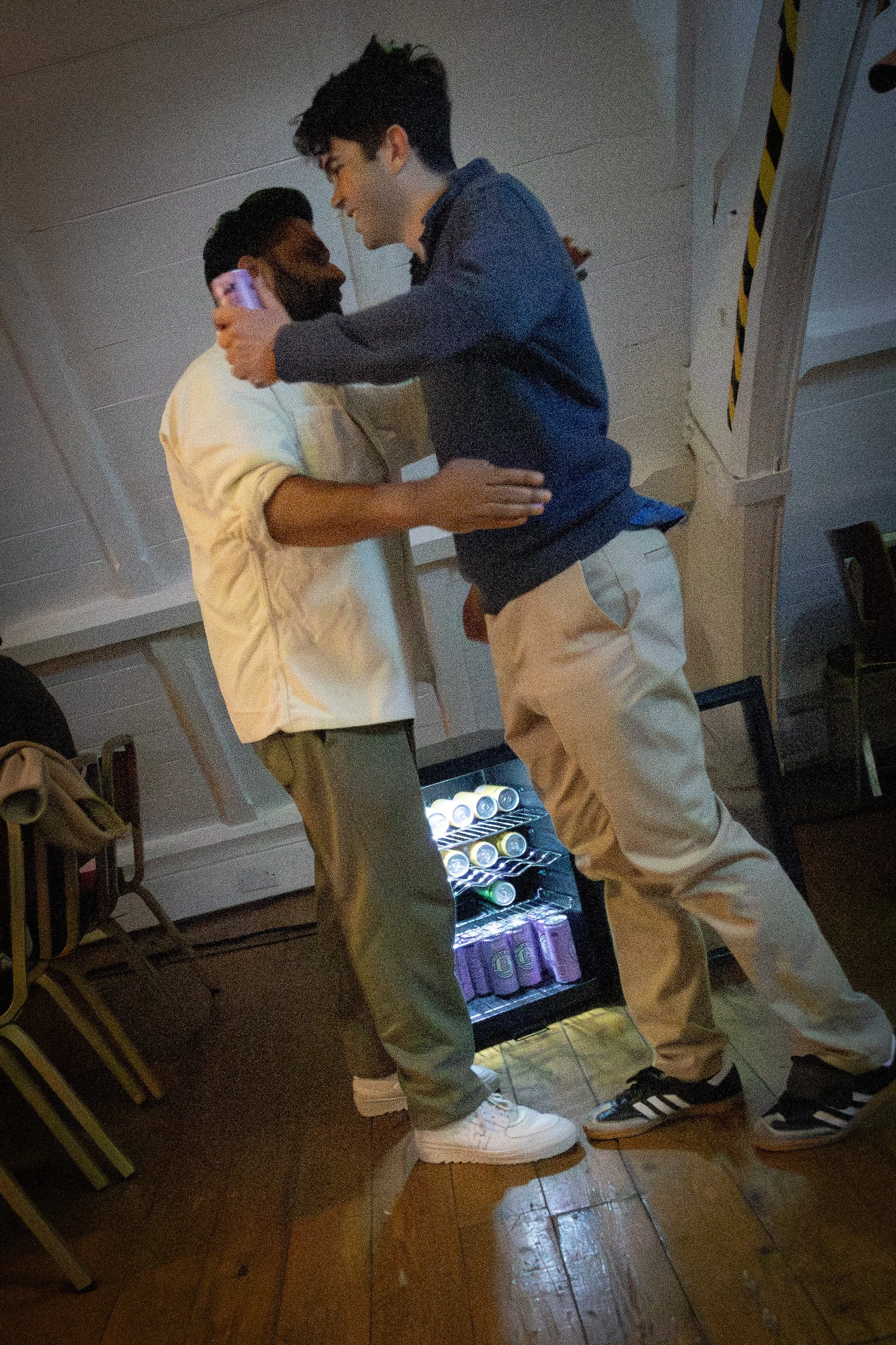 Two young men standing close, one holding a phone and the other with a hand on the other's waist, inside a room with a liquor refrigerator and several chairs.