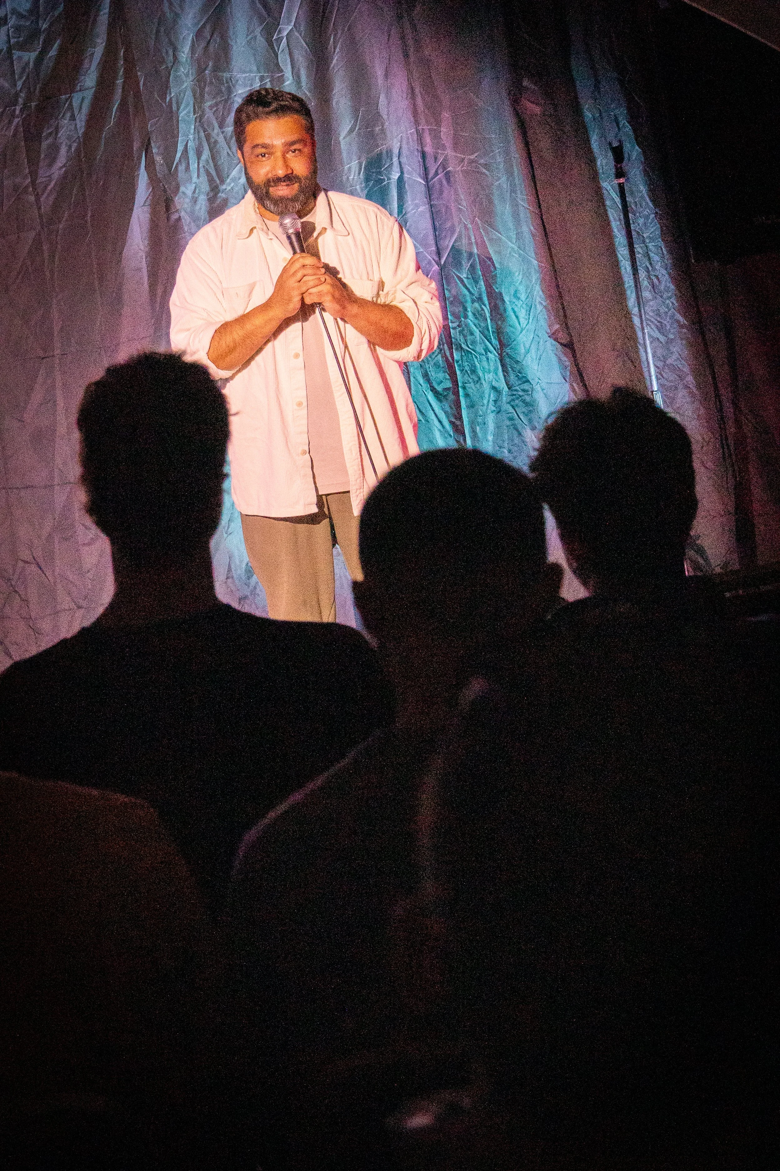 A man holding a microphone on stage with a crinkled dark blue curtain backdrop, facing the audience.