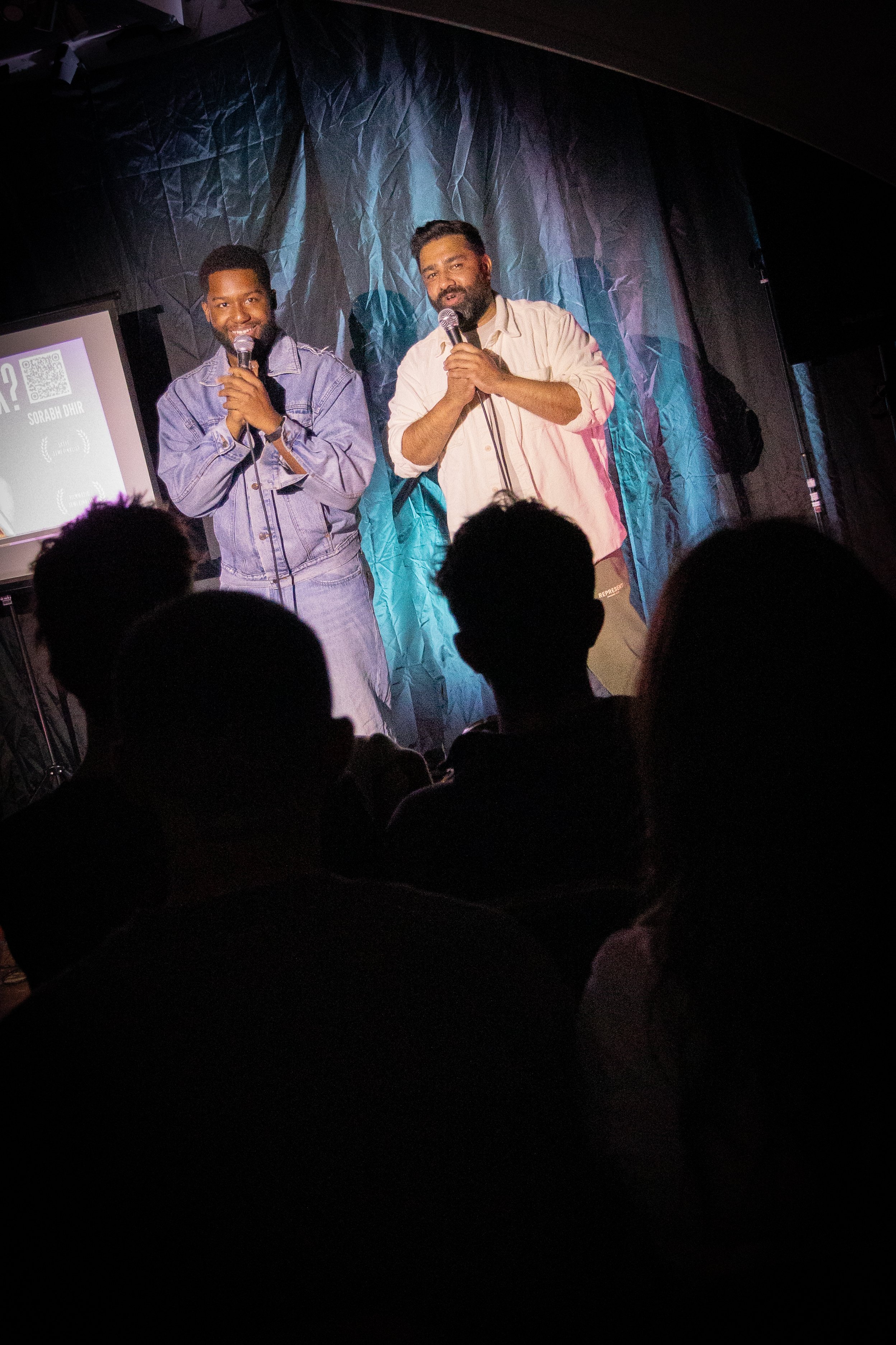 Two men on stage holding microphones, speaking to an audience, with a black curtain backdrop and a projection screen displaying text and a QR code.