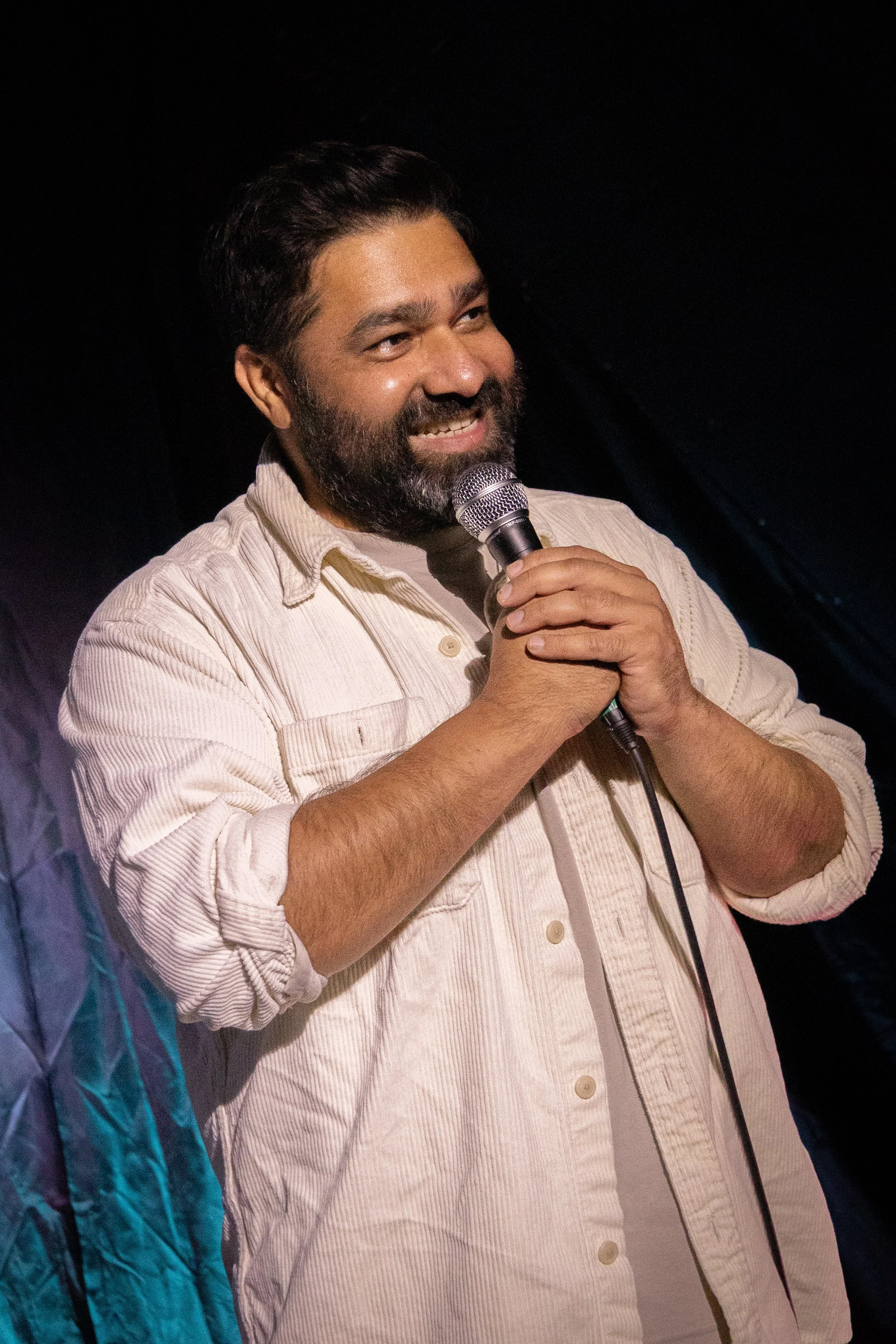 A man with a beard and mustache holding a microphone and smiling, wearing a light-colored, long-sleeve, button-up shirt.