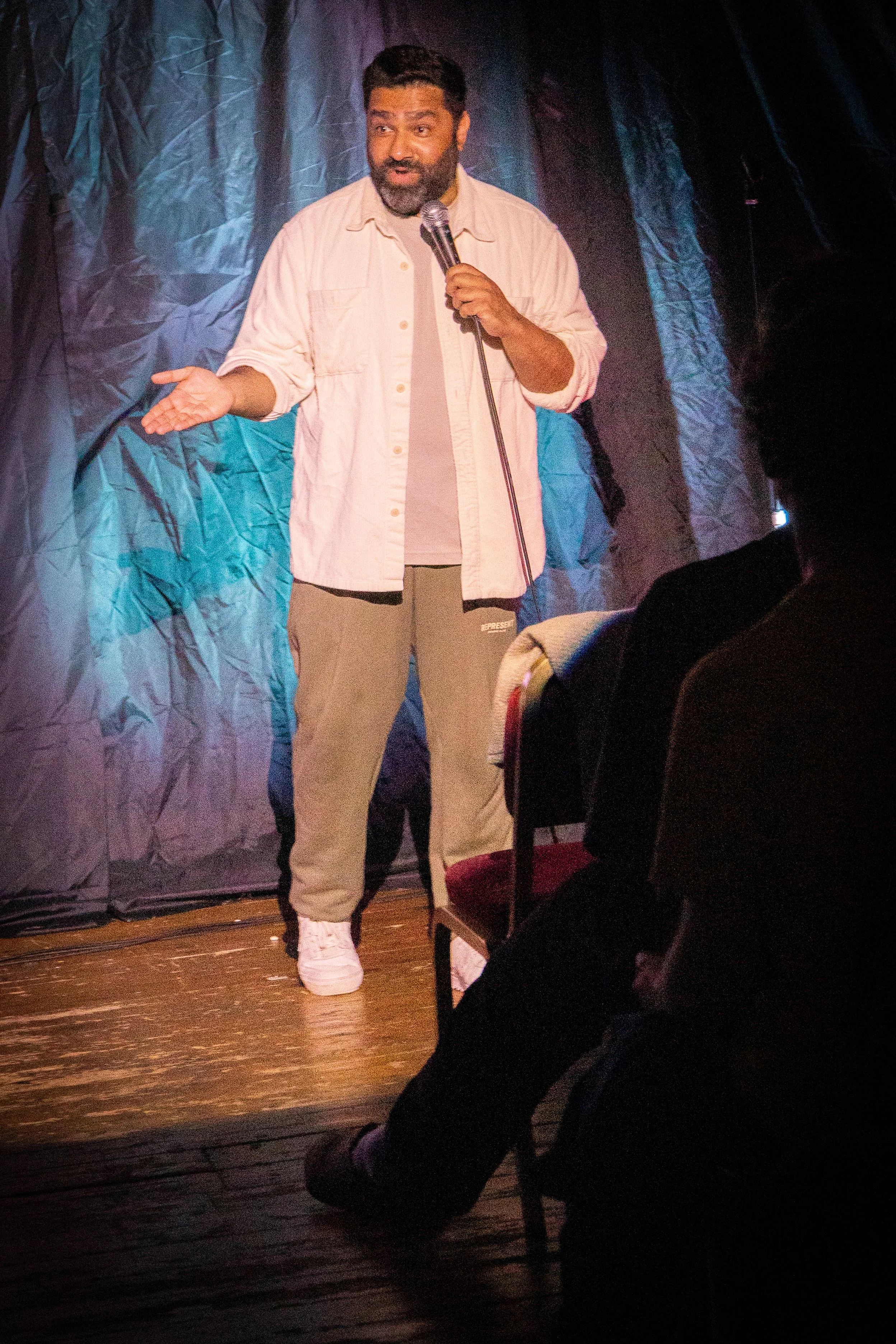 A man with a beard, wearing a light-colored jacket and beige pants, stands on stage holding a microphone with one hand and gesturing with the other. There is a dark, textured curtain behind him and an audience member's head is visible in the foregrou