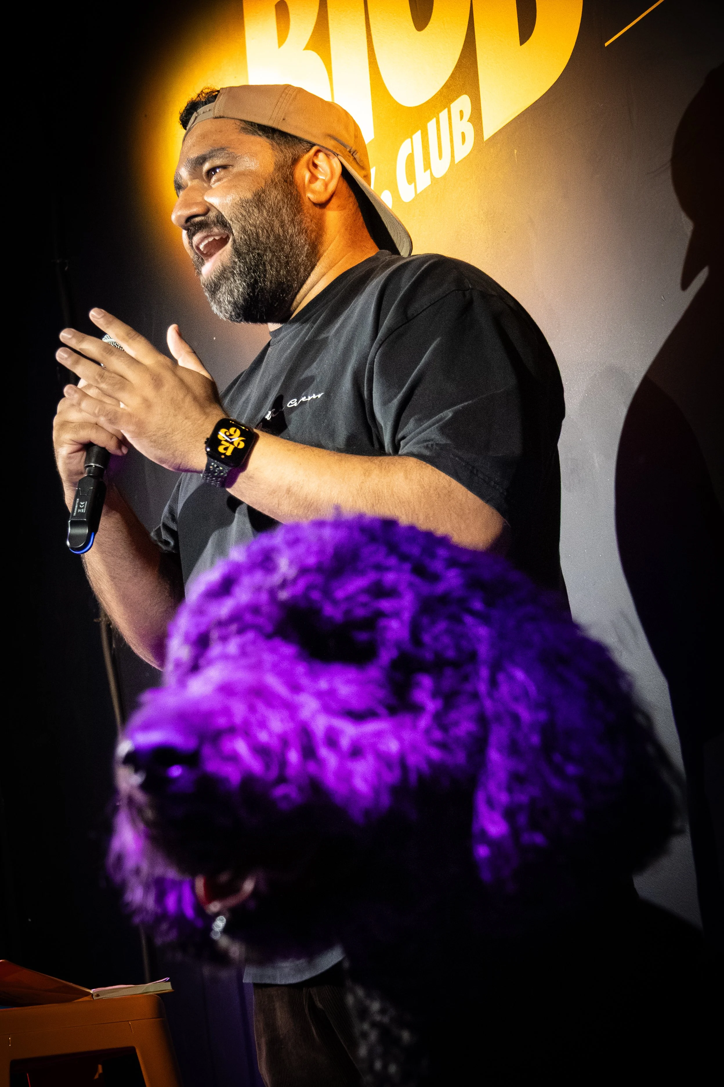 A man with a beard wearing a baseball cap backwards, speaking into a microphone on stage during a stand-up comedy performance at the Bood Room Comedy Club. The background features a yellow wall with the club's name in large white letters.