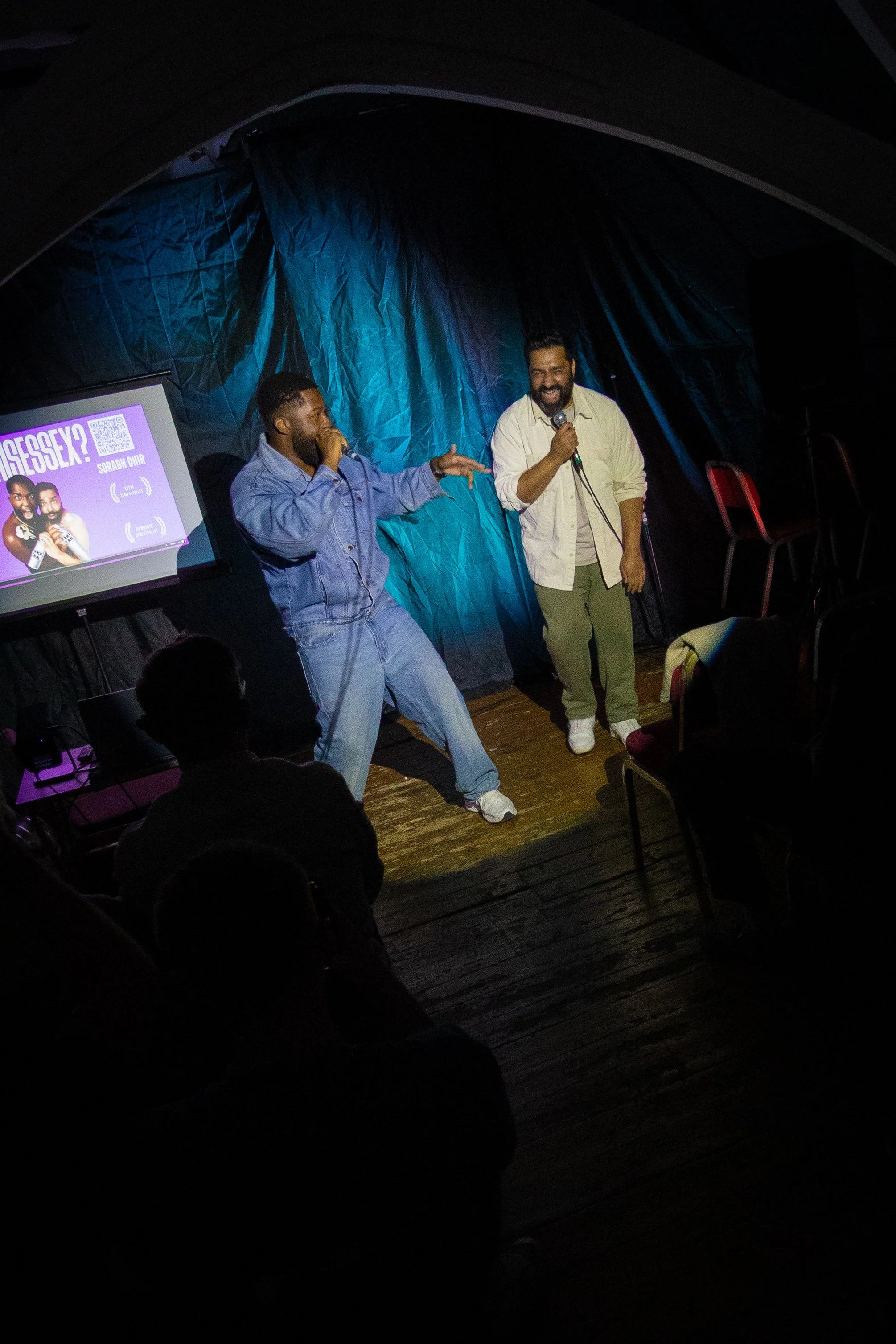 Two men are on stage, one holding a microphone and speaking, the other making a gesture, both smiling. There are chairs by the stage and a screen displaying a poster with photos of two women, text, and a QR code.