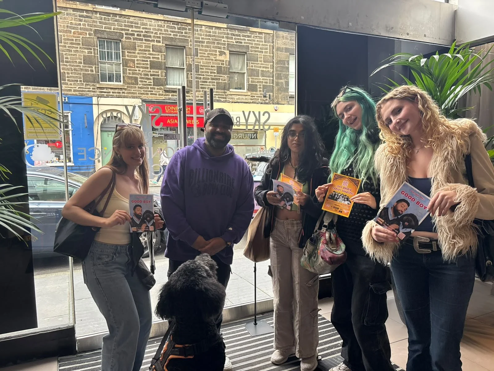 Group of five people and a dog standing inside a building near a glass door, holding promotional flyers and brochures, with a city street visible outside.