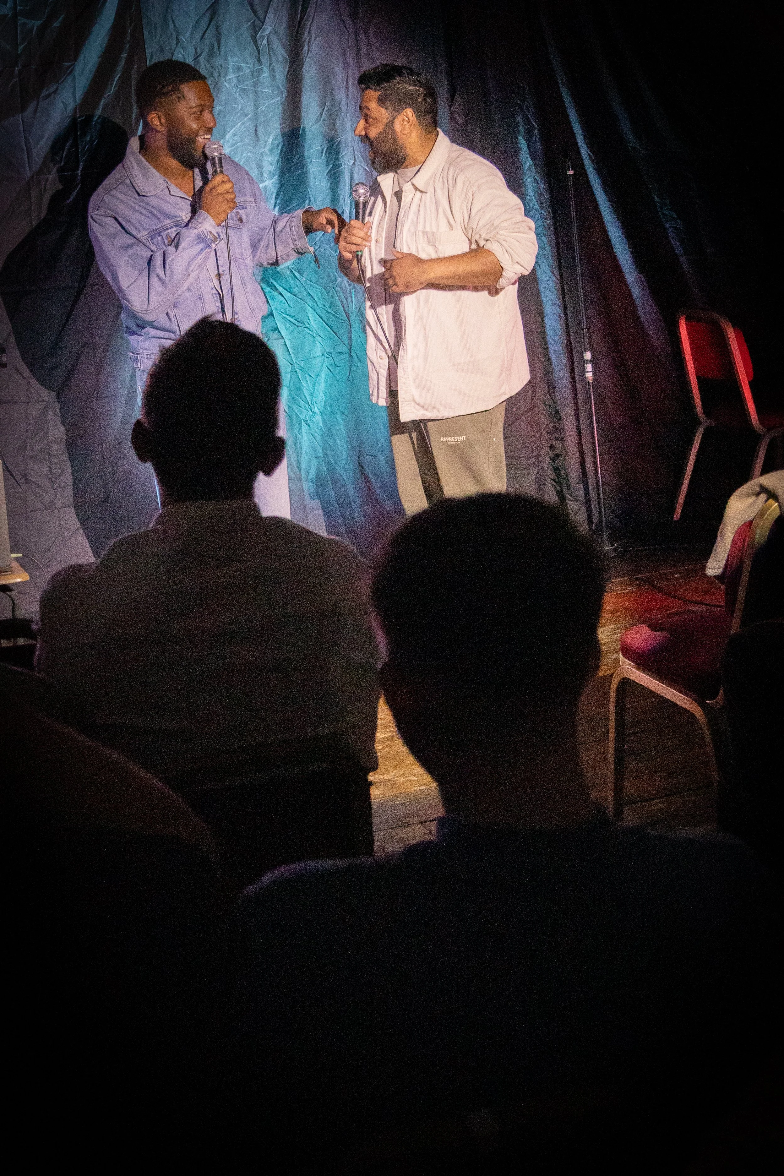 Two men are standing on a stage, engaging in a conversation or performance. One man on the left is holding a microphone and smiling, wearing a denim jacket. The other man on the right, also holding a microphone, is smiling and wearing a light-colored
