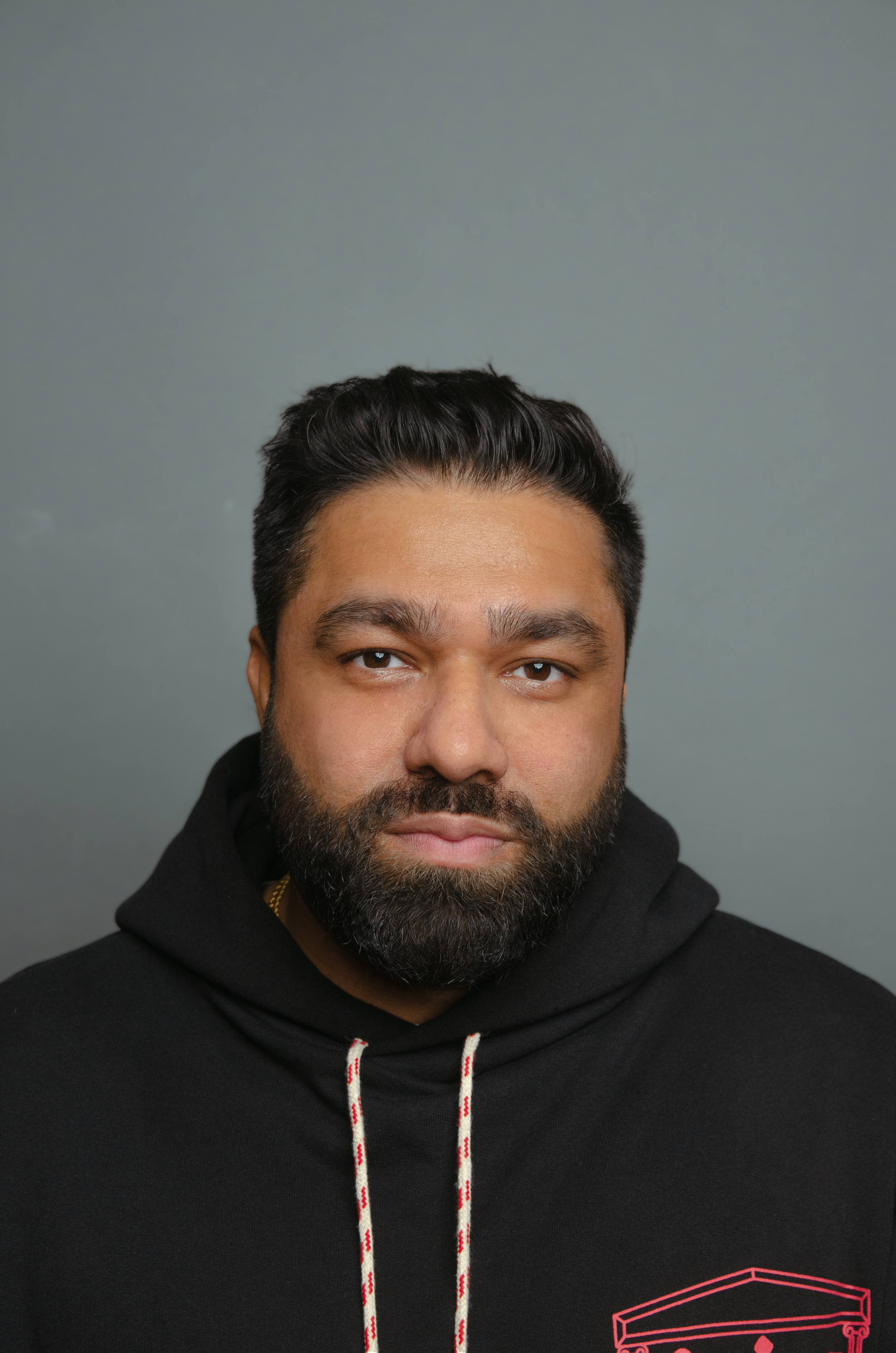 Close-up of a man's face with a beard and dark hair, wearing a black hoodie with a red graphic design, against a gray background.