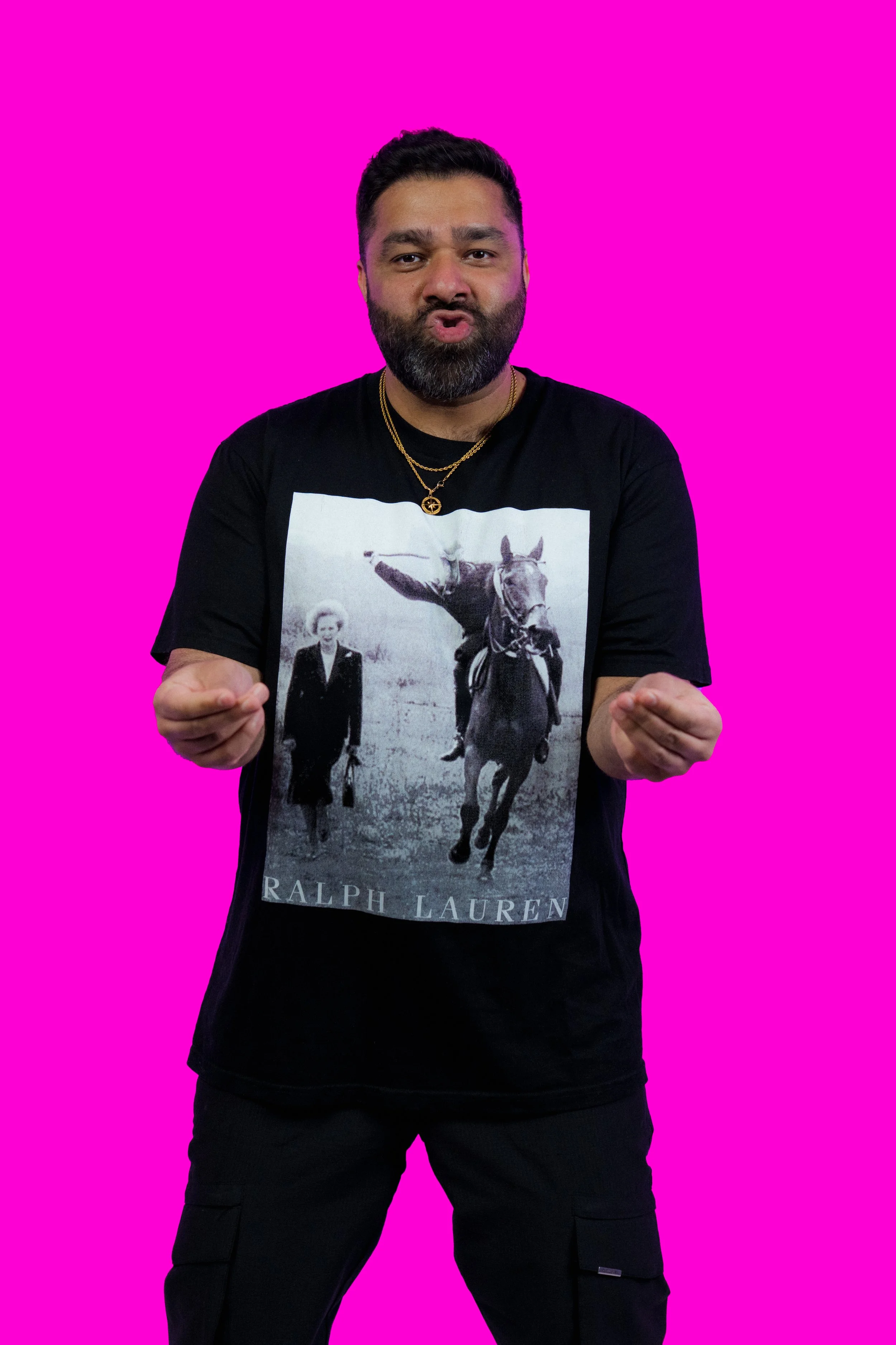 A man with dark hair and beard, wearing a black Ralph Lauren t-shirt with a vintage black-and-white photo of a woman and a horse, gold chain necklaces, standing against a bright pink background, making fists with both hands.