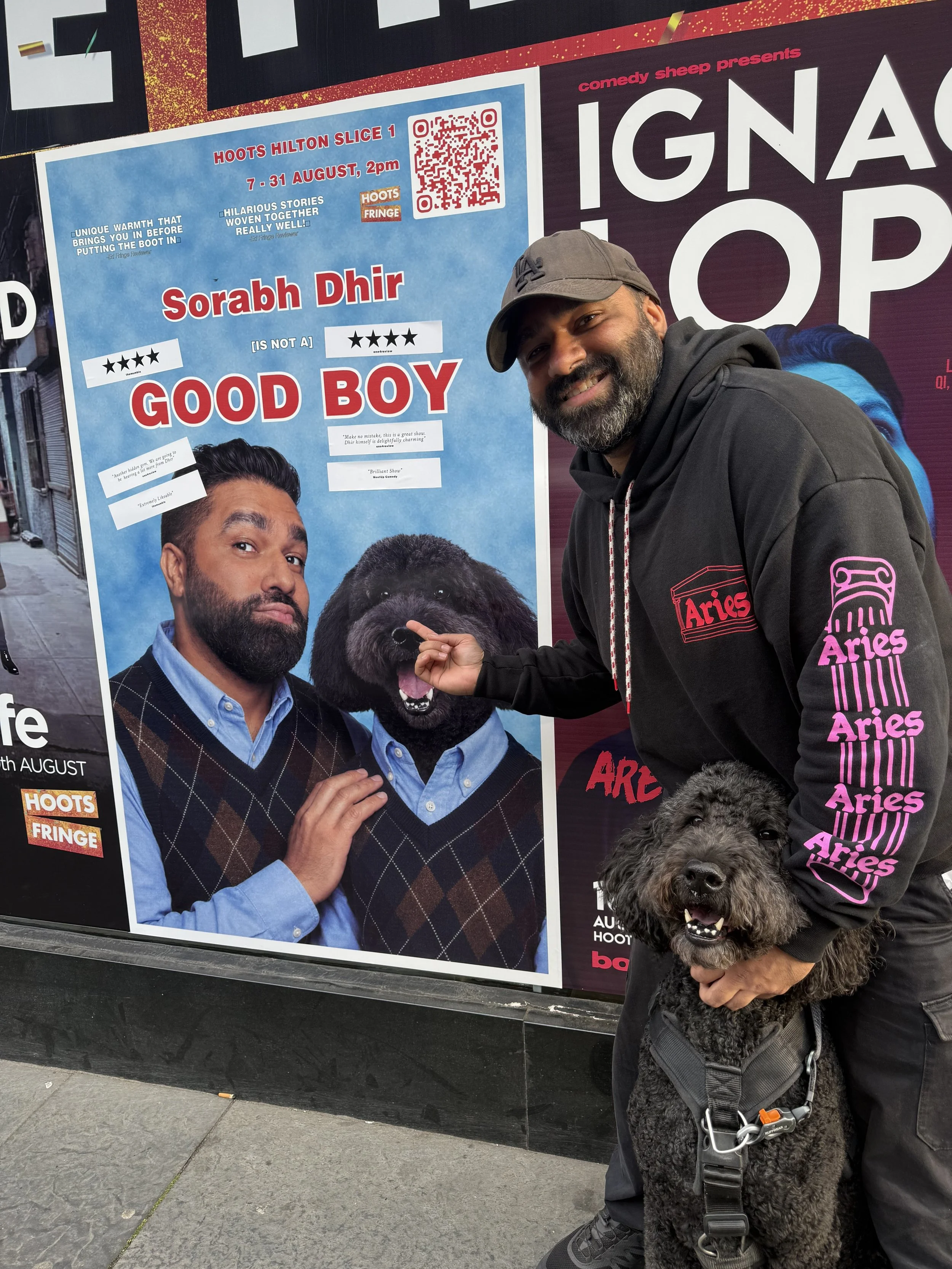 A man with a beard wearing a gray cap and a black hoodie holding a black curly dog with a harness next to a colorful event poster on the street.