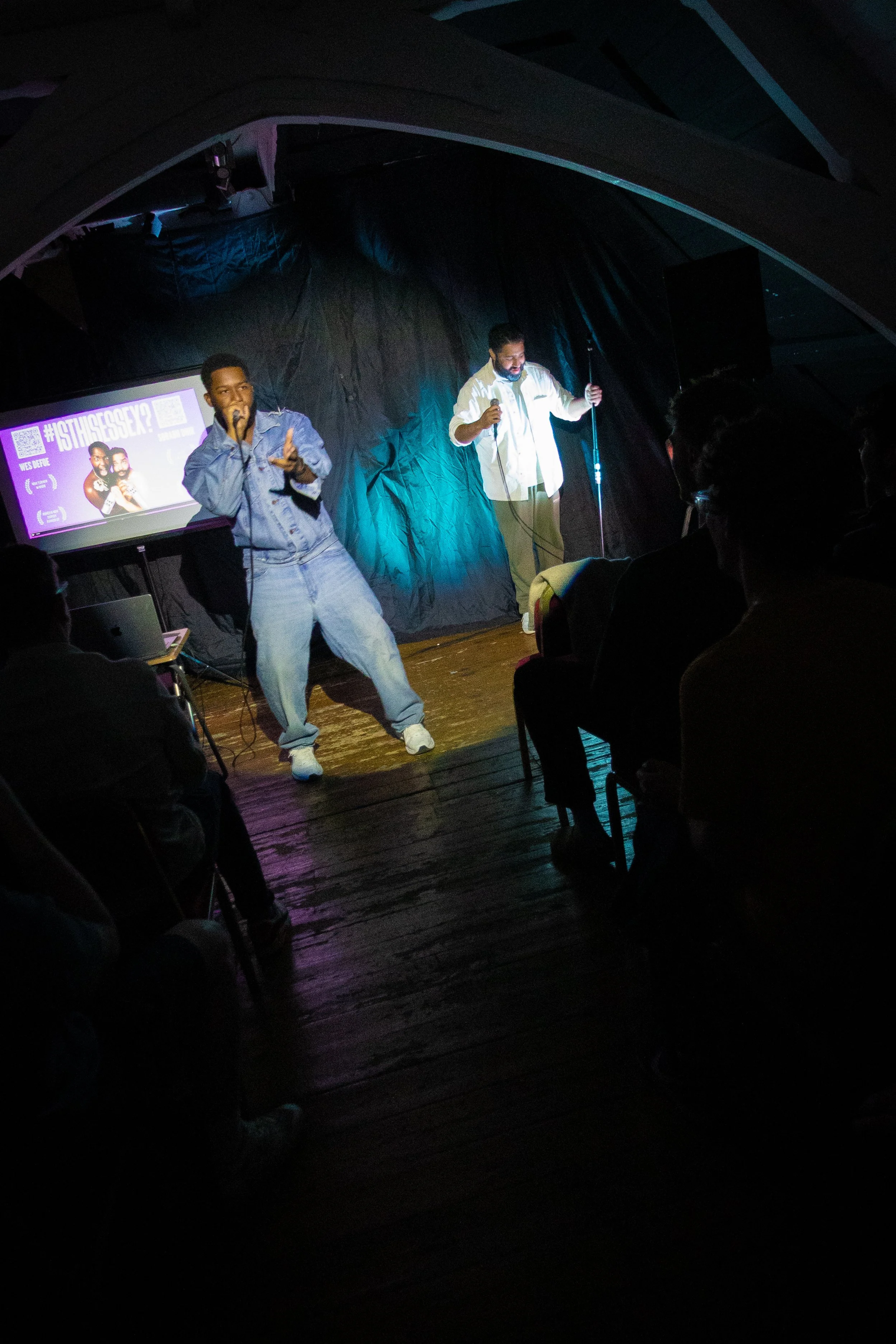 Two men performing on stage in a dark venue, one singing into a microphone and the other holding a microphone stand, with a projected poster behind them.