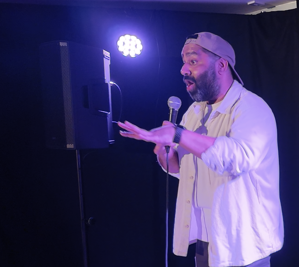 Male comedian wearing a backward cap, performing with a microphone on stage with purple lighting.