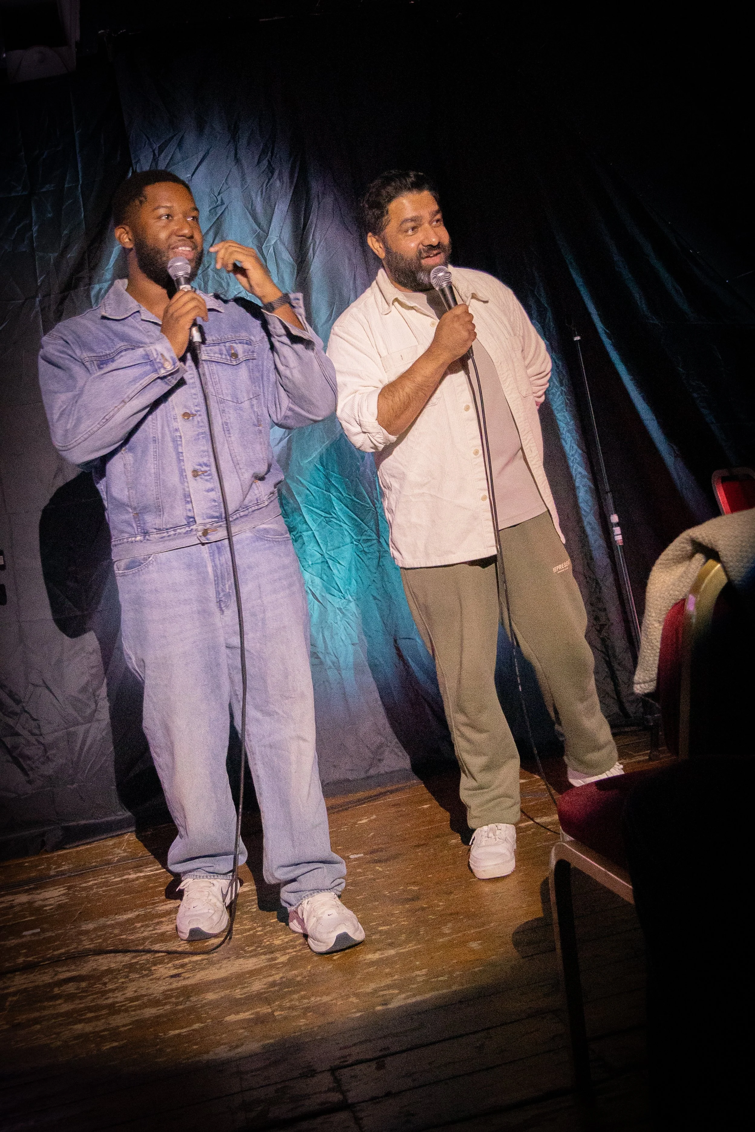 Two men are standing on a wooden stage, holding microphones, and speaking. The man on the left is wearing a denim jacket and jeans, and the man on the right is wearing a light-colored shirt and pants. There is a dark curtain in the background, and so