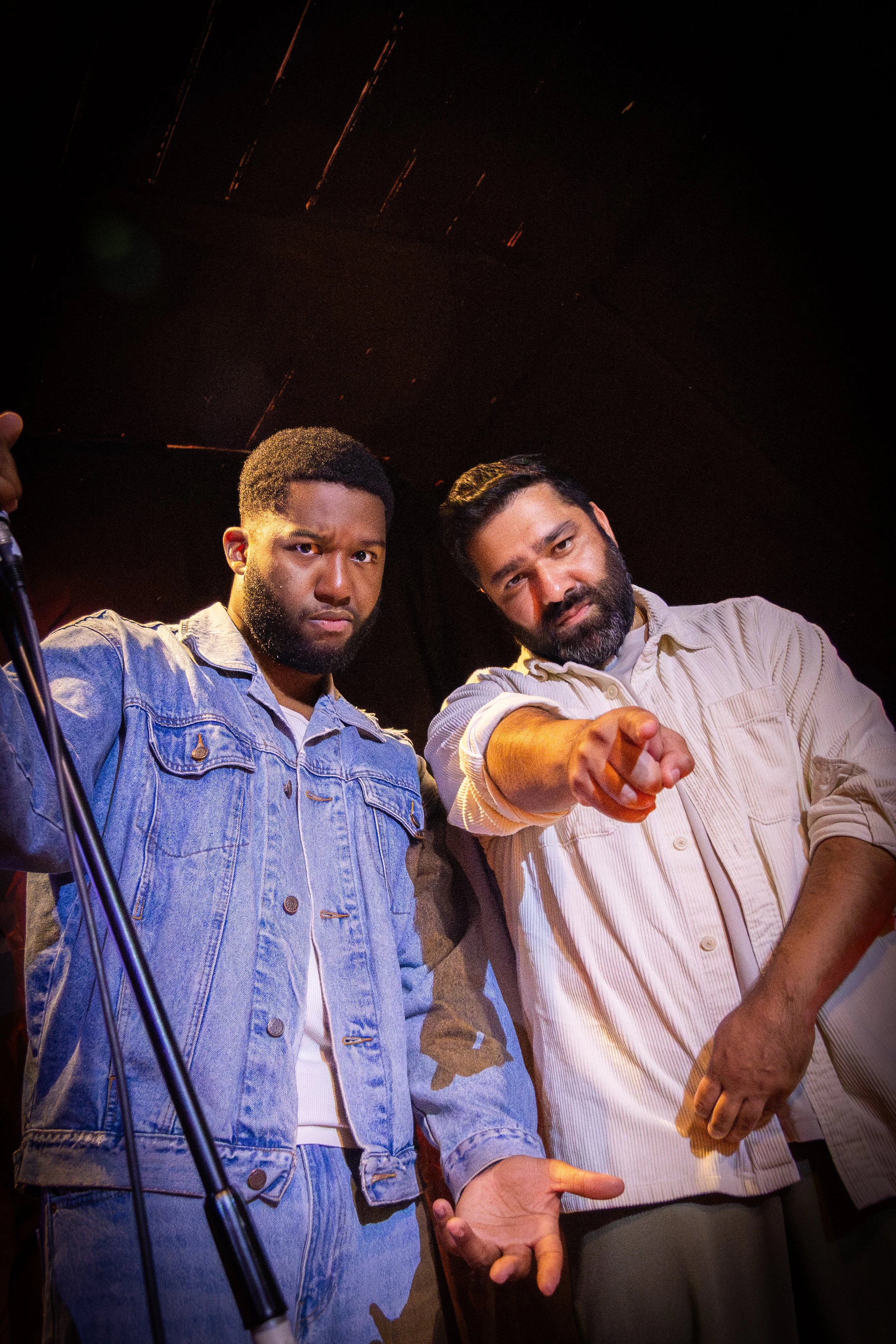 Two men, one wearing a denim jacket and the other in a light-colored shirt, facing the camera under dim lighting, with a microphone stand partially visible on the left side of the image.