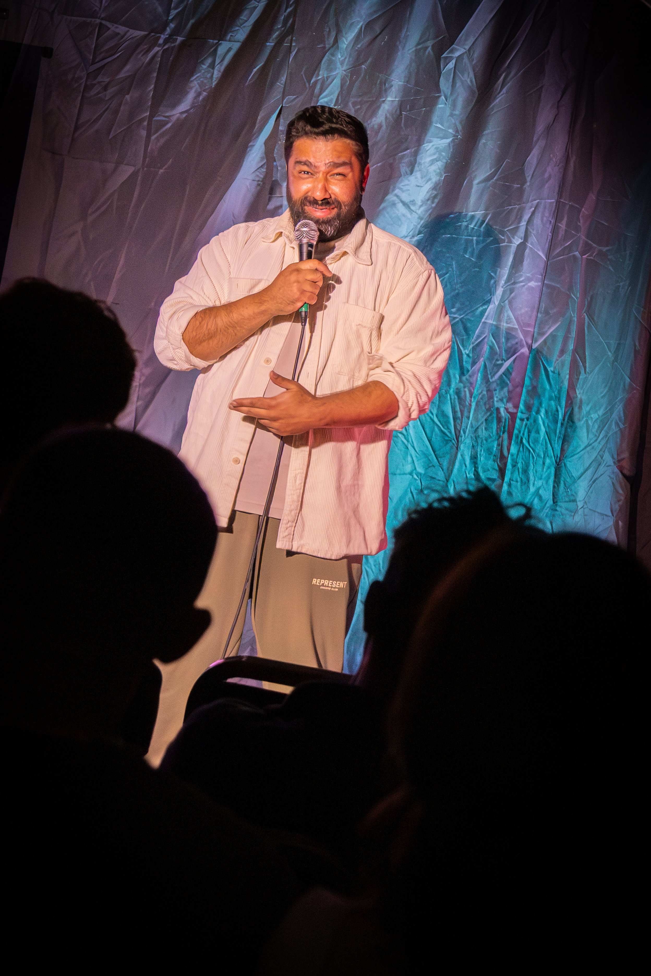 A man with a beard and mustache holding a microphone on stage, with a crinkled blue and purple backdrop, facing an audience.
