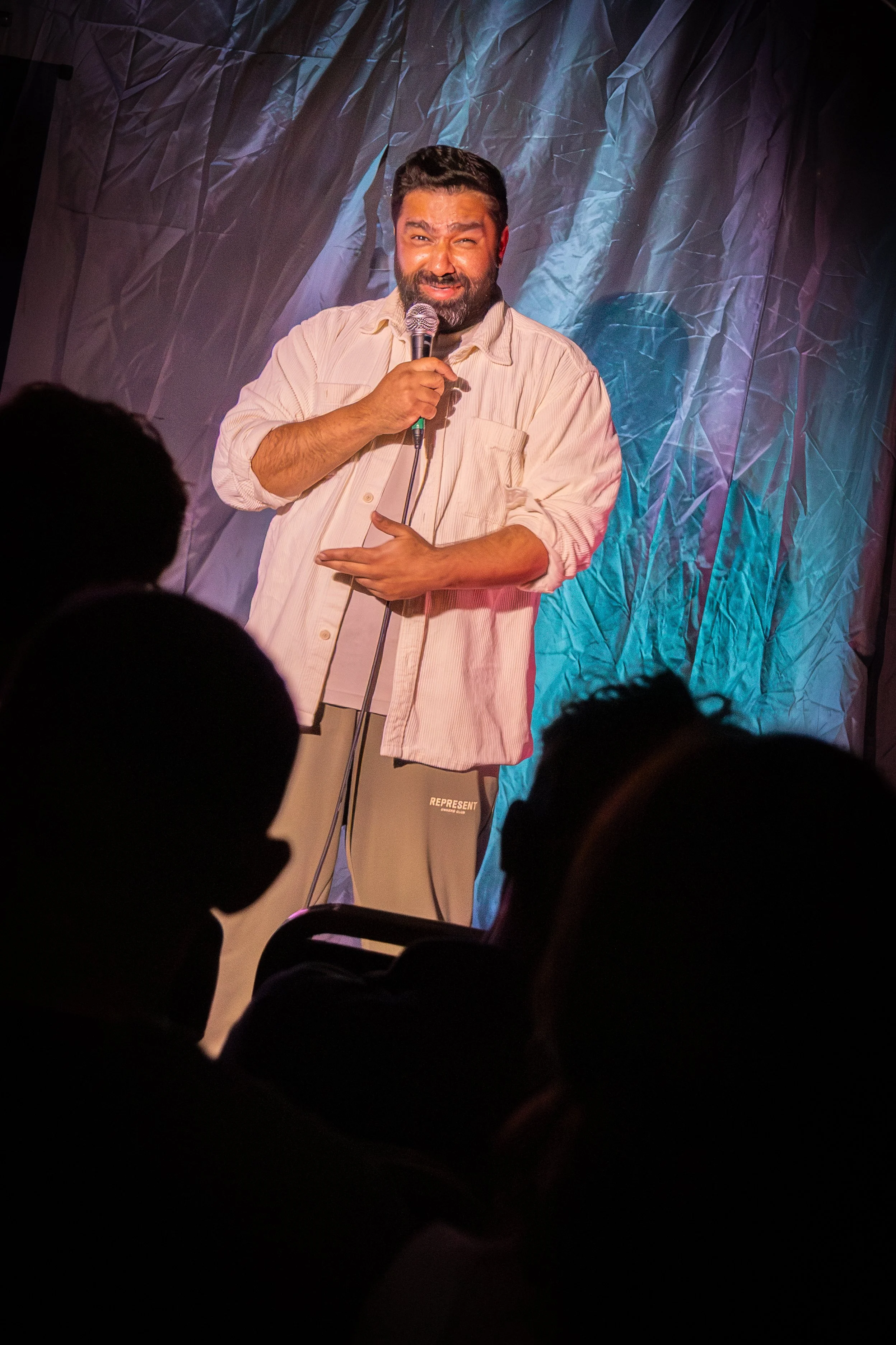 A man with a beard and dark hair holding a microphone on stage, wearing a light-colored shirt and beige pants, with a crinkled blue and purple backdrop behind him, during a performance or comedy show.