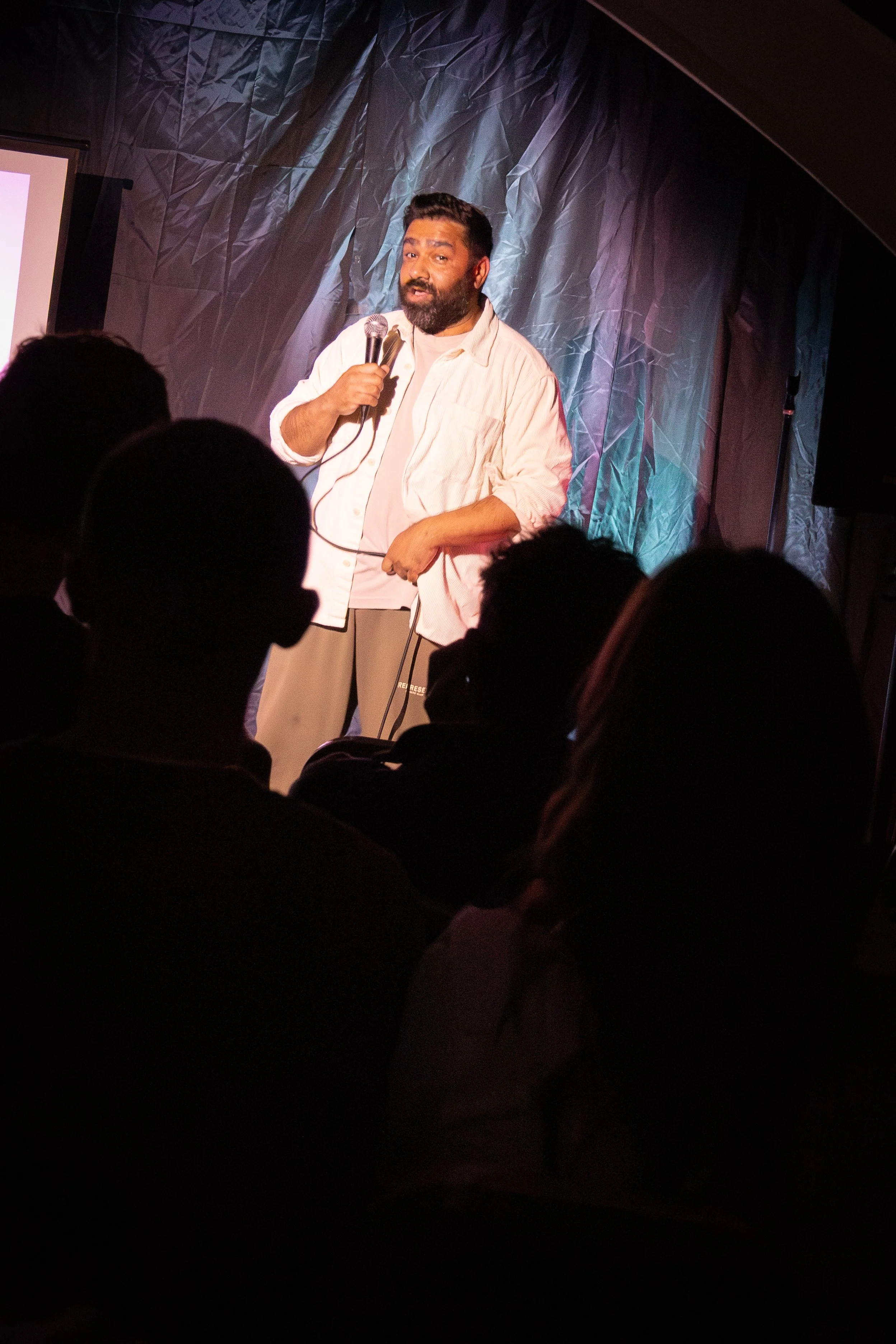 A man with a beard wearing a light-colored shirt holding a microphone, speaking on stage with a dark, textured backdrop, and an audience in front.