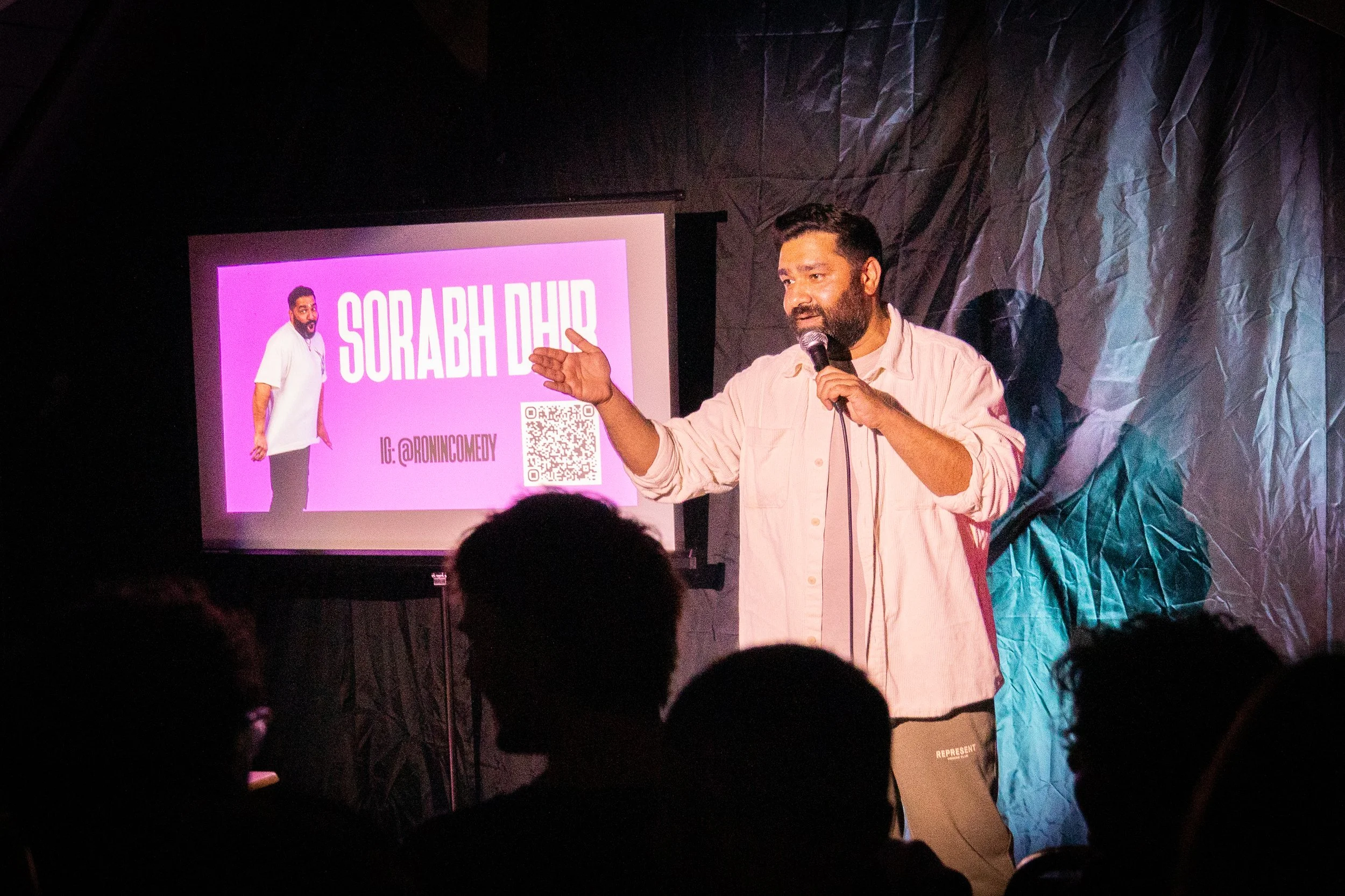A man with dark hair and beard holding a microphone and gesturing with his left hand, speaking on stage at a comedy event. There is a large screen behind him displaying his name 'Sorab Dahi' in white text on a pink background along with his Instagram