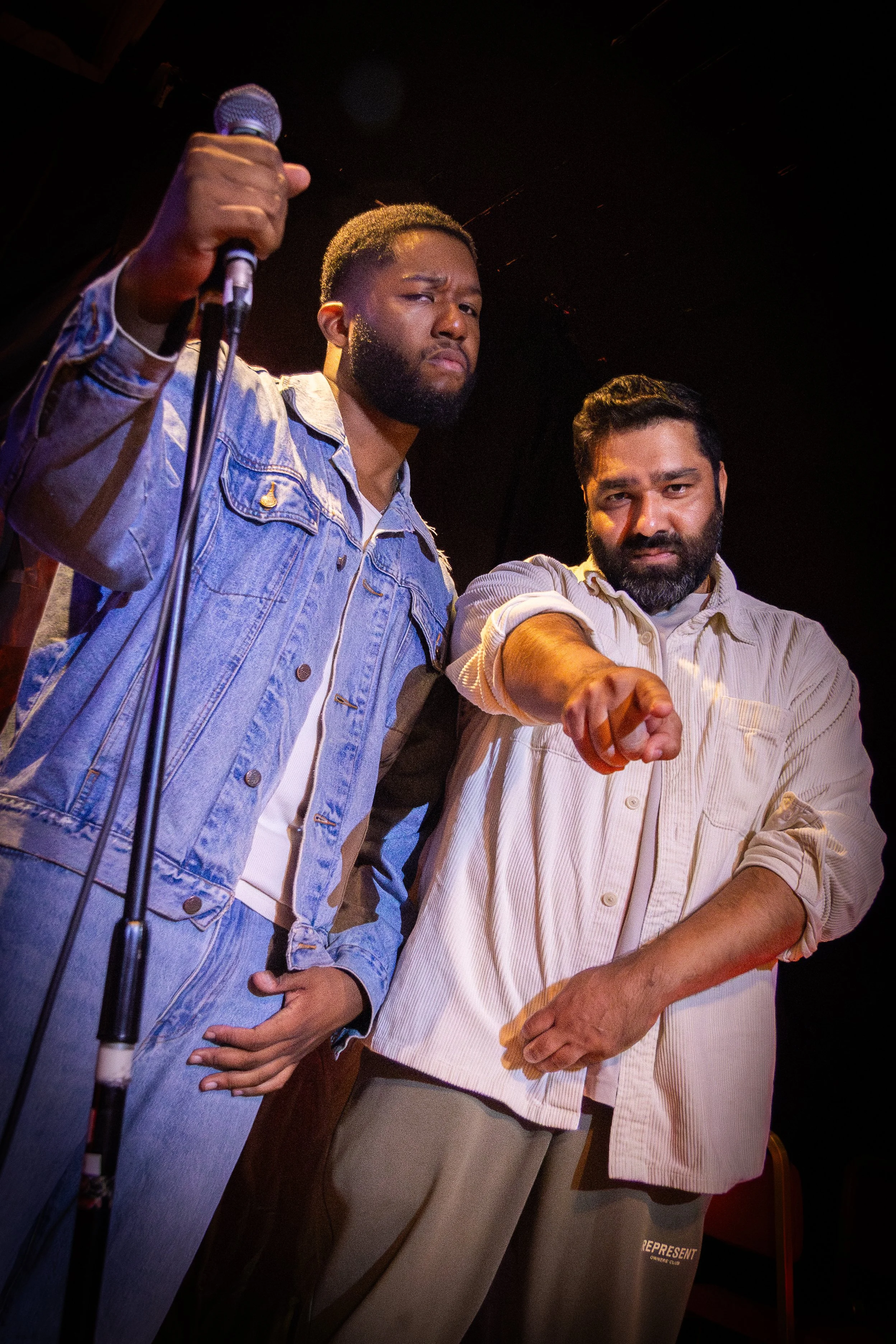 Two men standing in front of a microphone, one holding the microphone and both pointing forward with serious expressions.