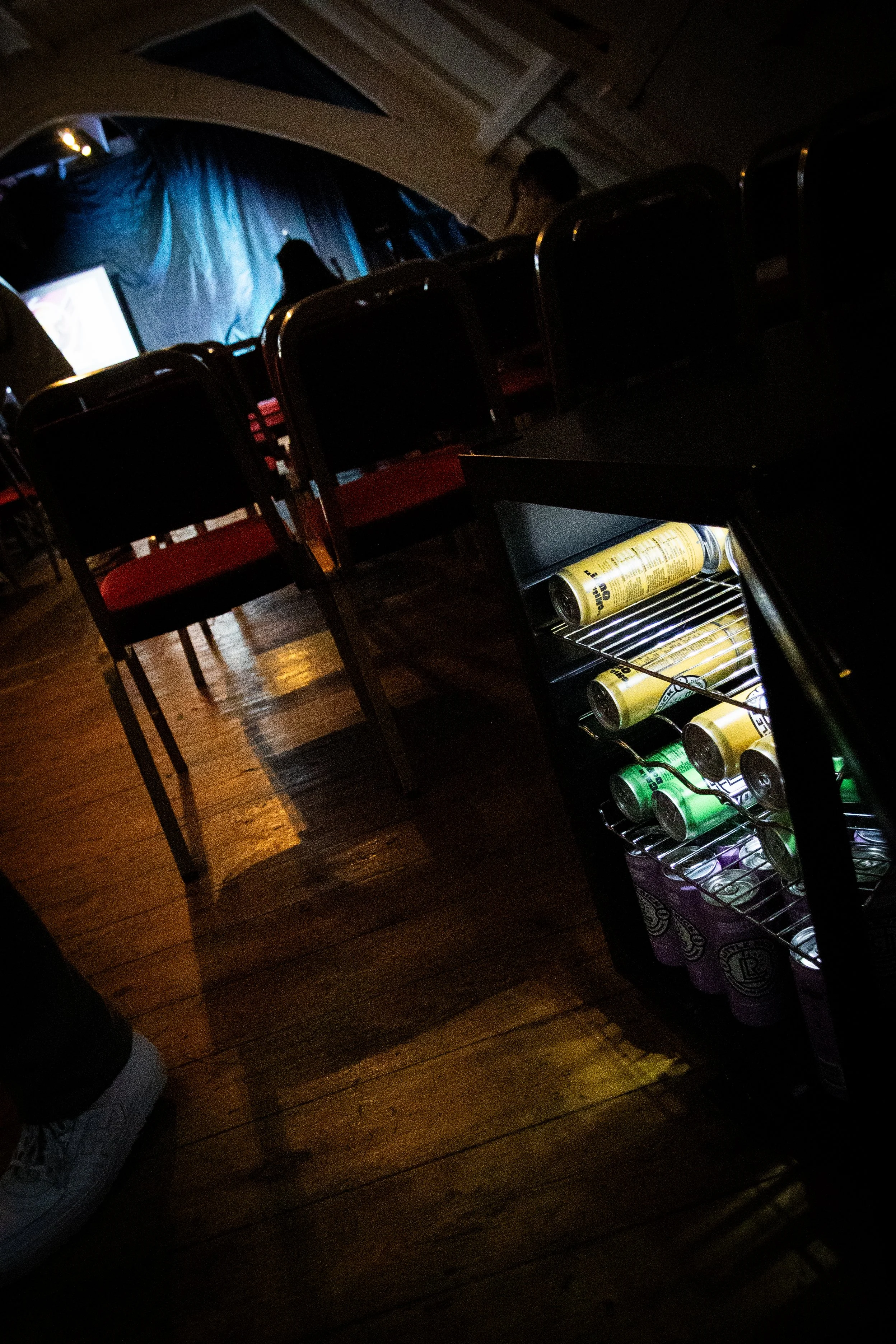 Dark image of a room with a small refrigerator filled with canned drinks, wooden flooring, chairs, a blue curtain, and people sitting and standing near a stage or screen, with a person’s head visible at the bottom left.