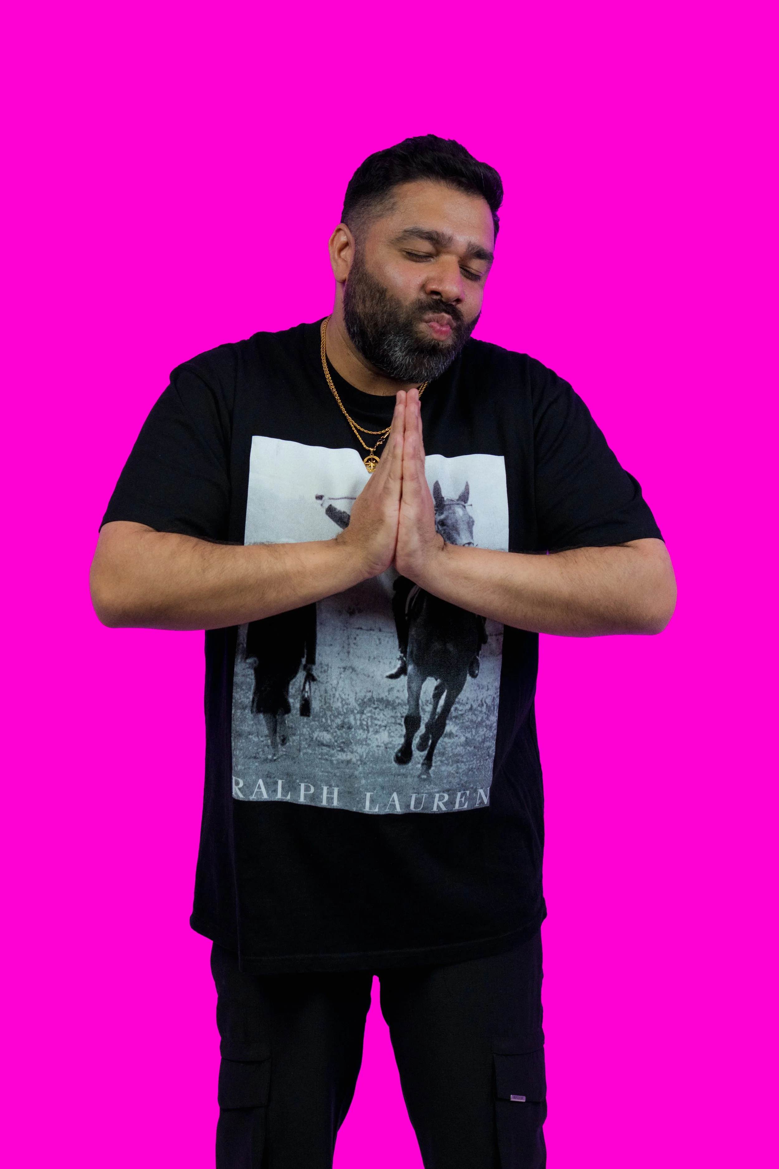 A man with dark hair and a beard, wearing a Ralph Lauren T-shirt with a horse graphic, black cargo pants, and gold necklaces, is standing against a bright pink background with his eyes closed and hands pressed together in a prayer gesture.
