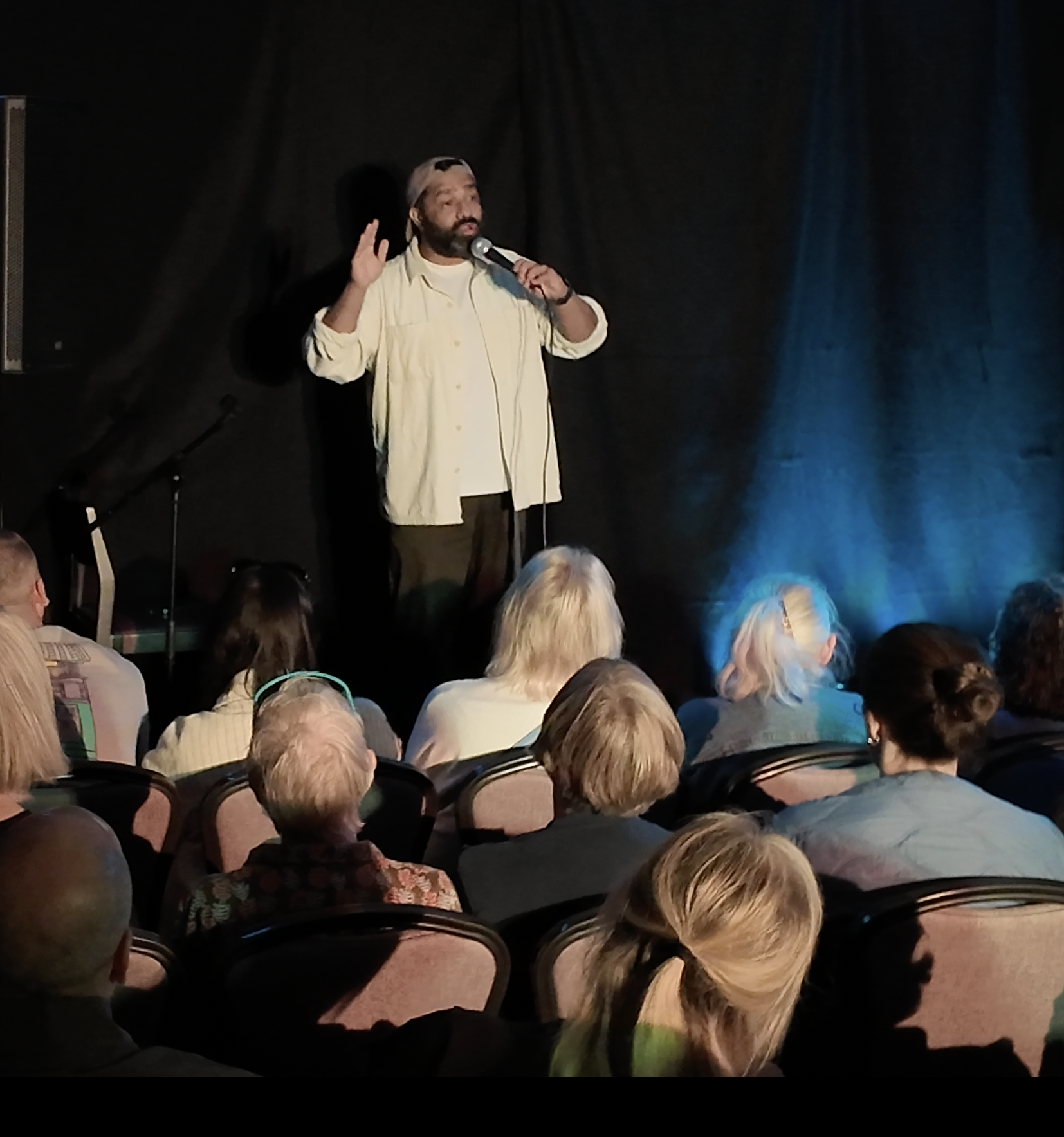 A man with a beard and a gray cap, wearing a light yellow shirt, is speaking into a microphone on stage. He has one hand raised, gesturing as he talks. The audience seated in front of him appears to be attentively listening. The stage is black with b