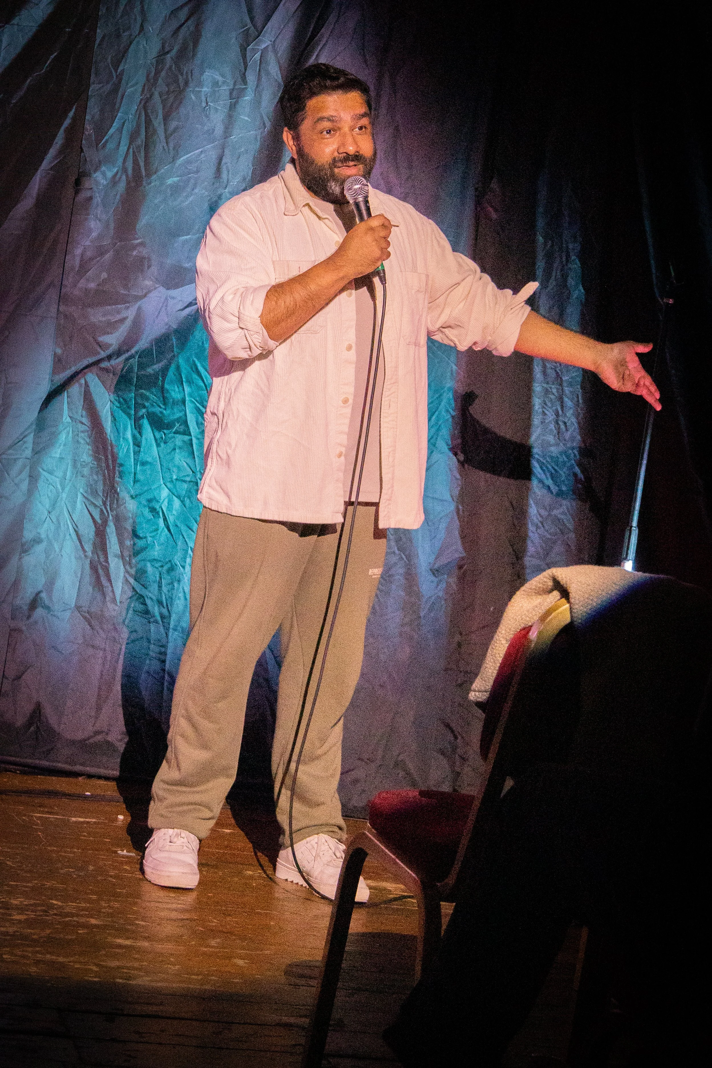 A man with a beard and dark hair, wearing a beige shirt, beige pants, and white sneakers, holding a microphone and speaking on stage with a dark, textured backdrop.