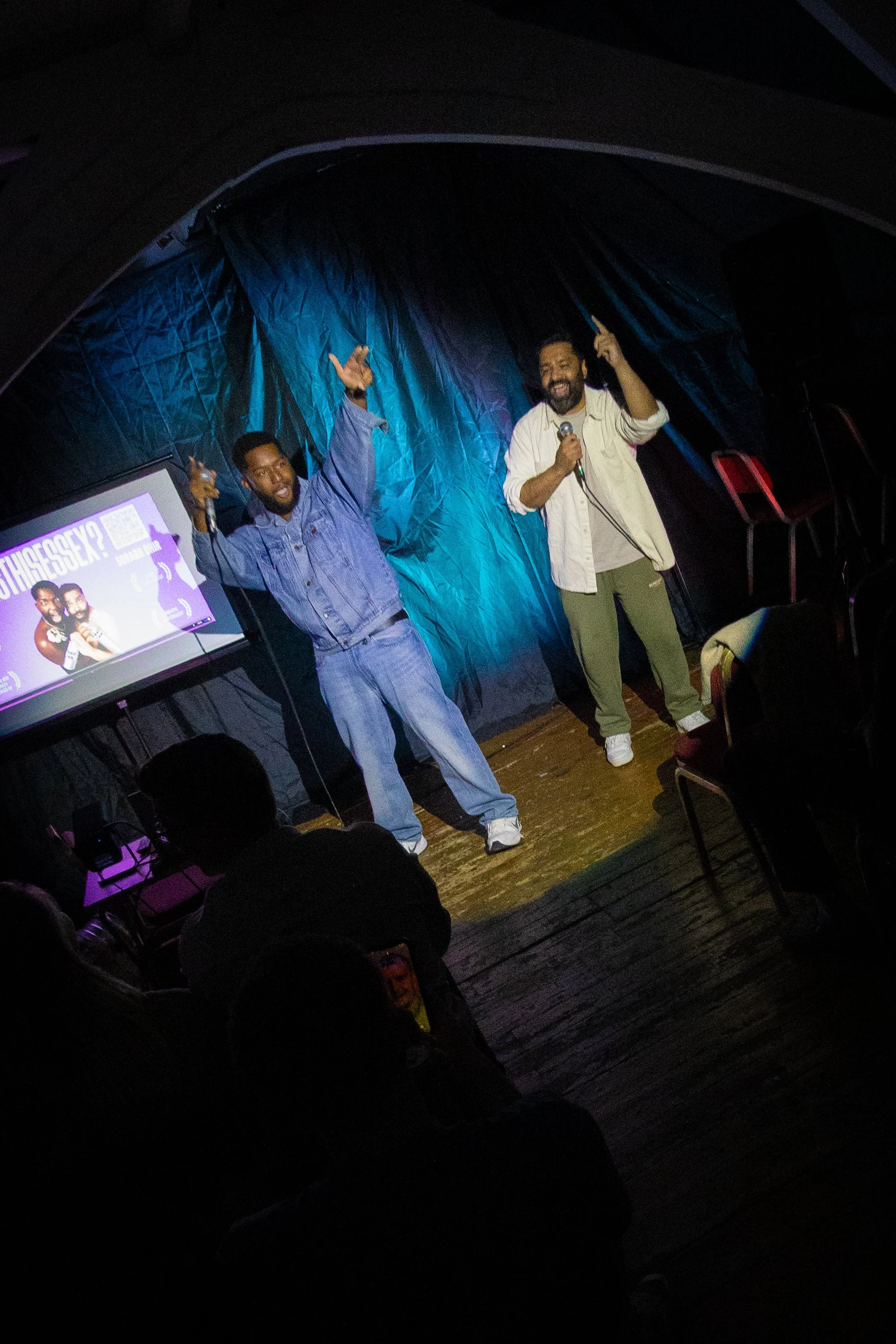 Two men on stage, one holding a microphone, raising their hands and smiling, with a blue curtain backdrop and a presentation slide in the background.