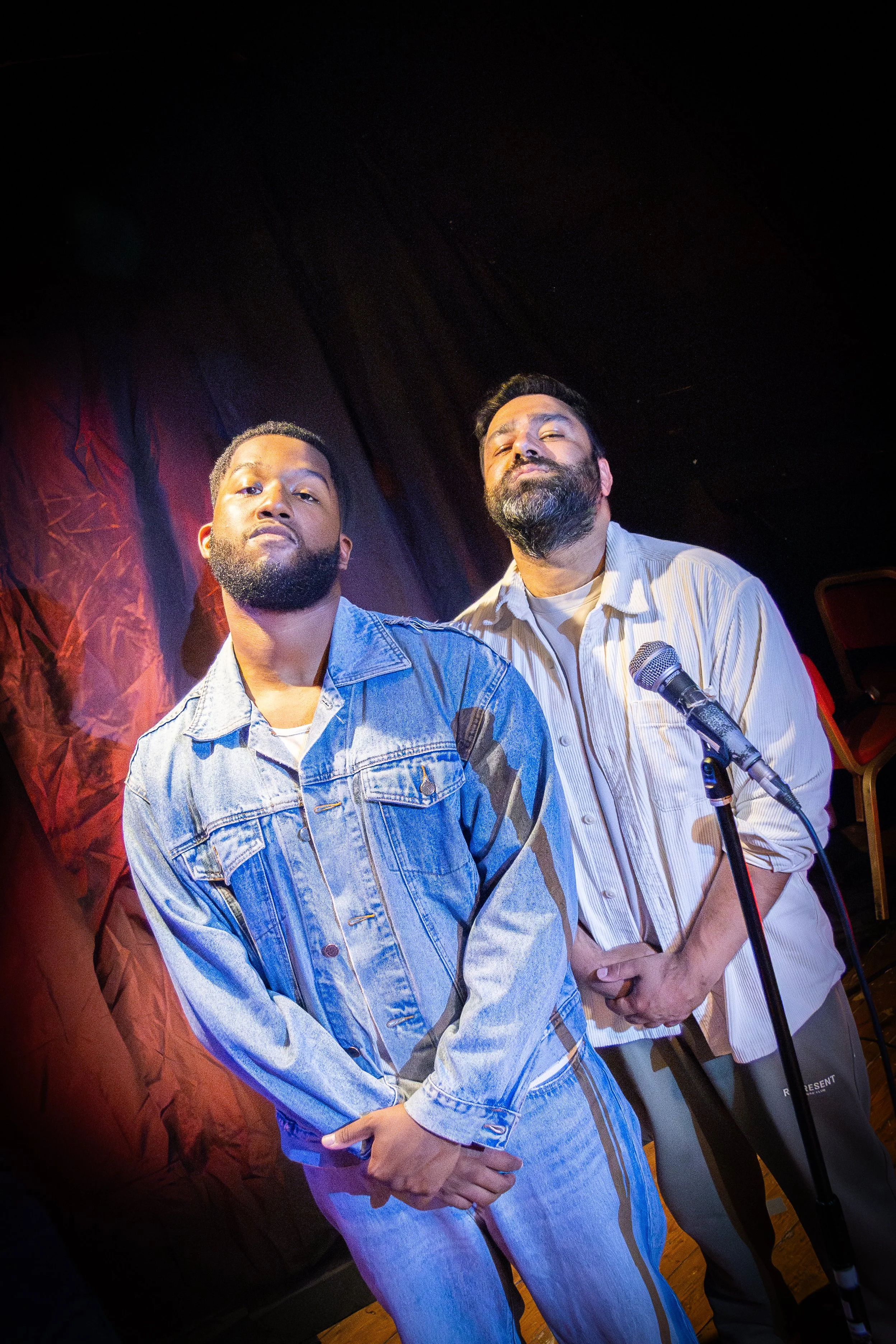 Two men on a stage in front of a dark backdrop, one holding a microphone, both posing confidently for a photo.