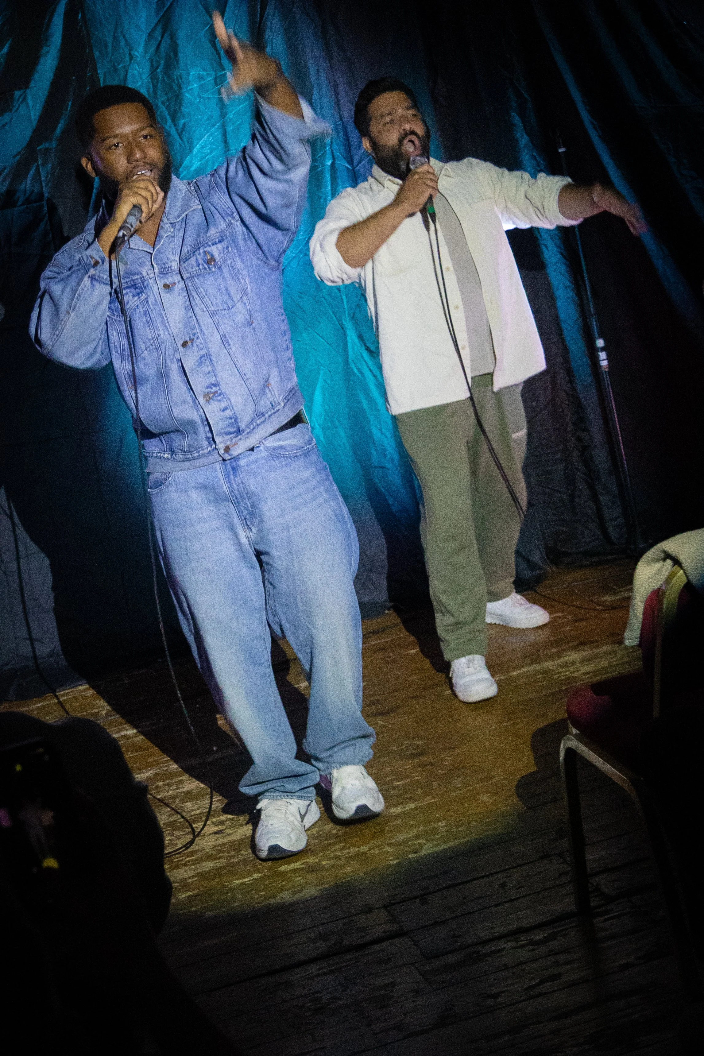 Two men are on a stage, singing or performing. One man is wearing a blue denim jacket and jeans, holding a microphone with his left hand and raising his right hand. The other man is wearing a white shirt and green pants, also holding a microphone and