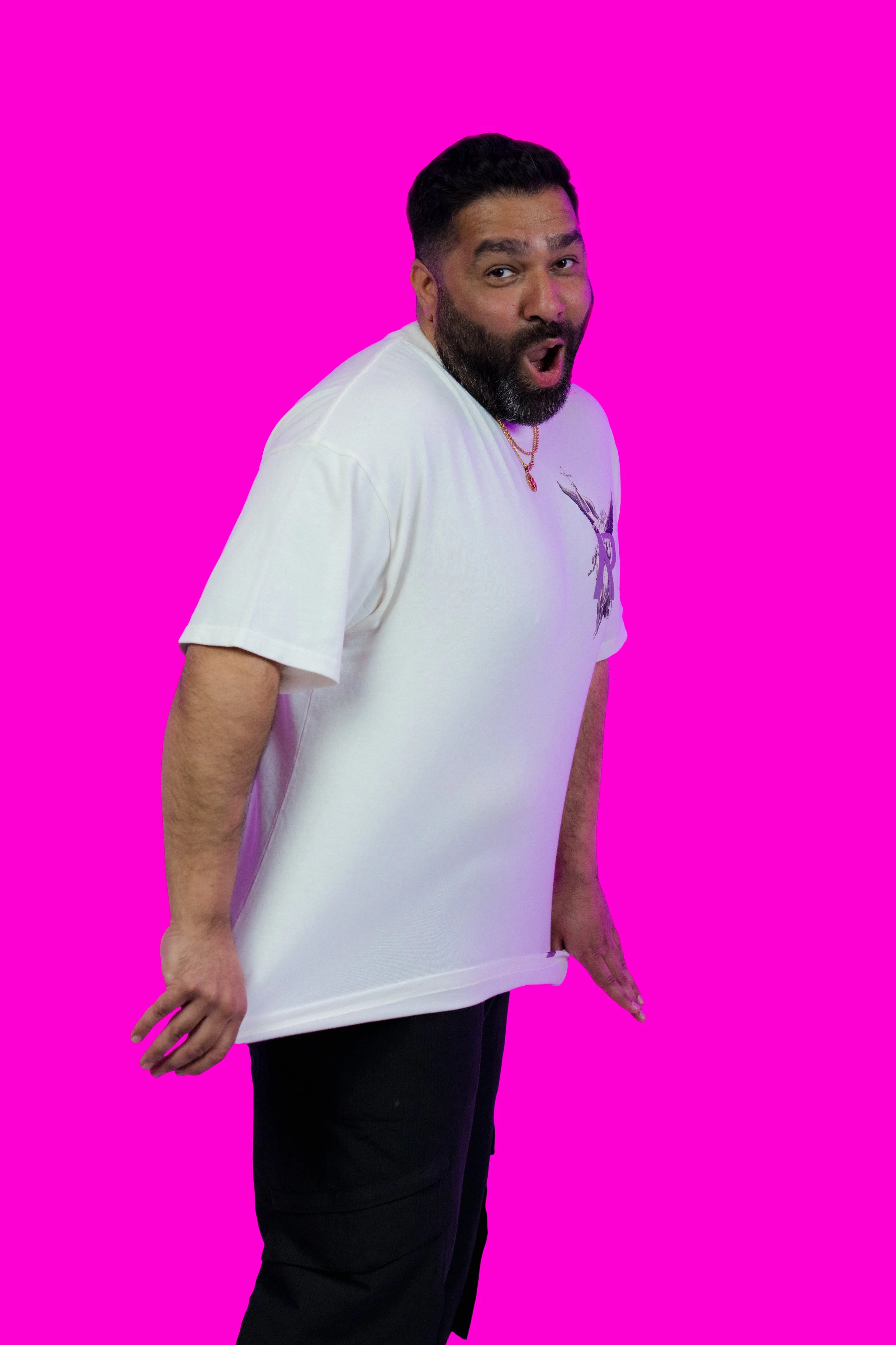 A man with a beard and short black hair wearing a white T-shirt and black pants, striking a pose against a bright pink background.
