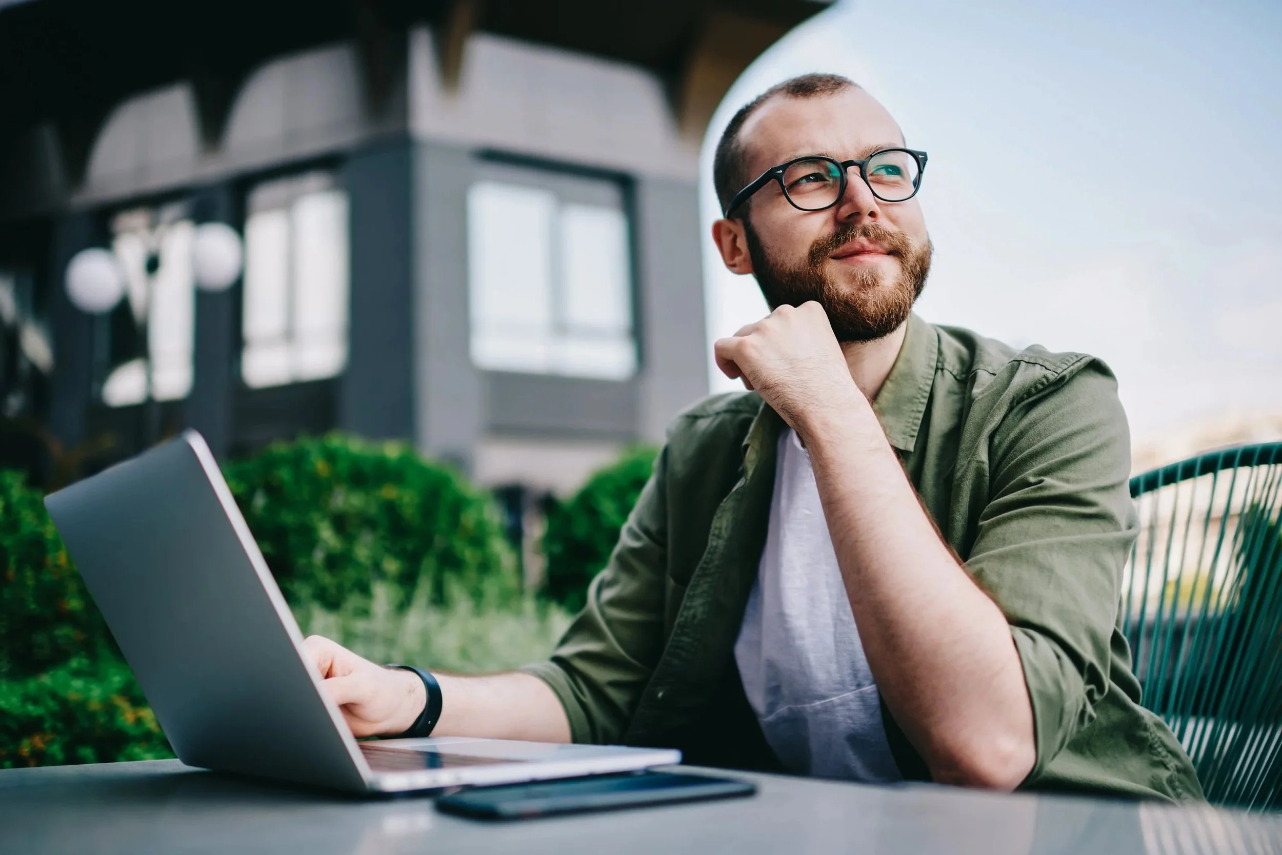 A man with glasses and a beard sitting at an outdoor table with a laptop, smiling with his hand near his neck, during daytime.