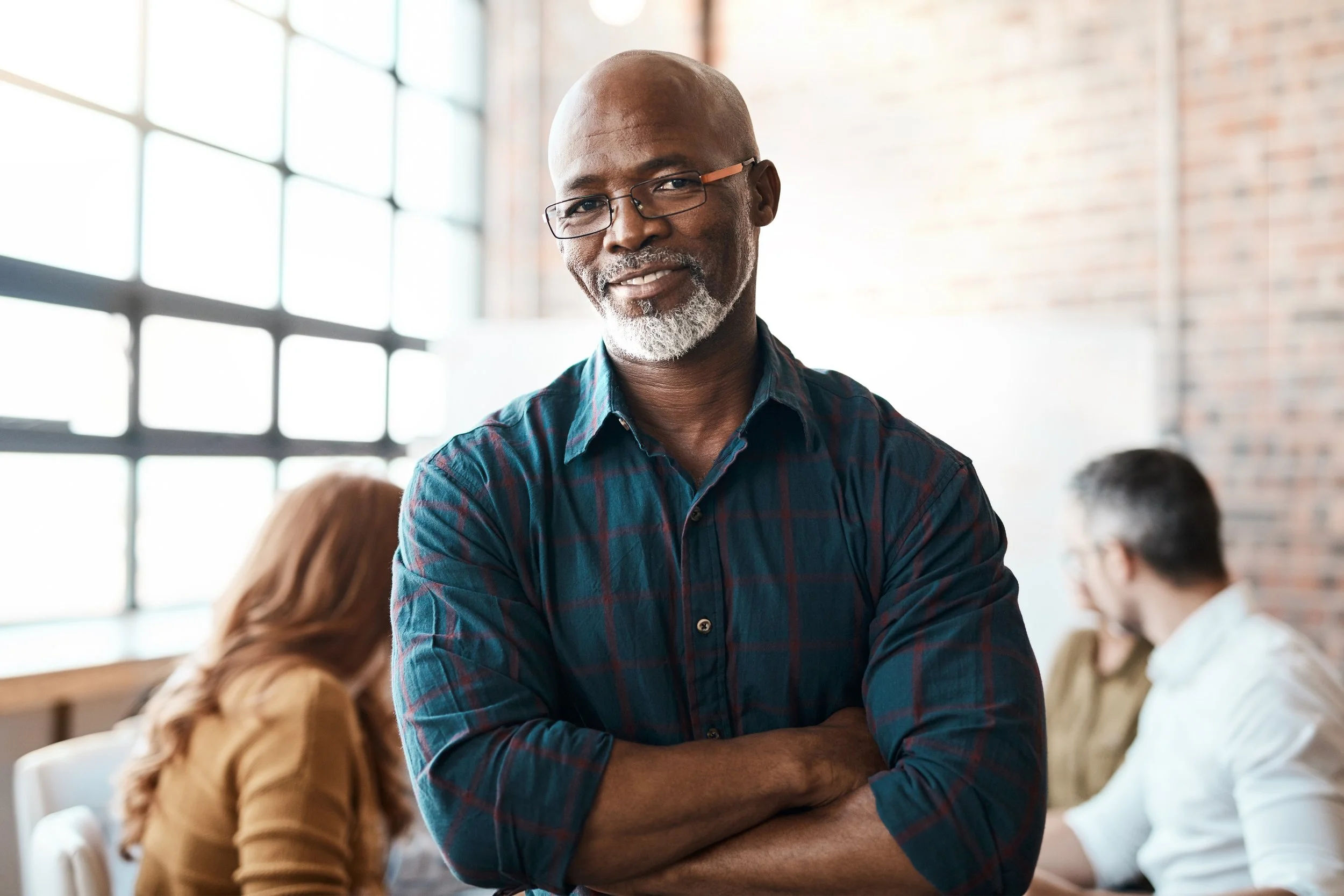 A confident middle-aged African American man with glasses and a gray beard wearing a blue plaid shirt, standing in a bright, modern office space with a brick wall and large windows, smiling and relaxing with his arms crossed.
