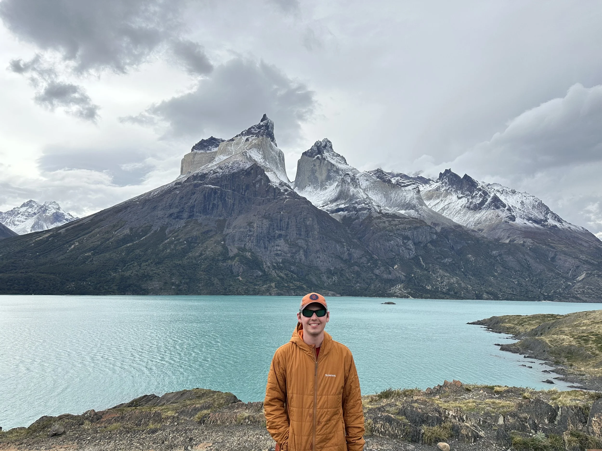 A person wearing sunglasses, an orange jacket, and a orange cap standing in front of a large turquoise lake with snow-capped mountains in the background under a cloudy sky.