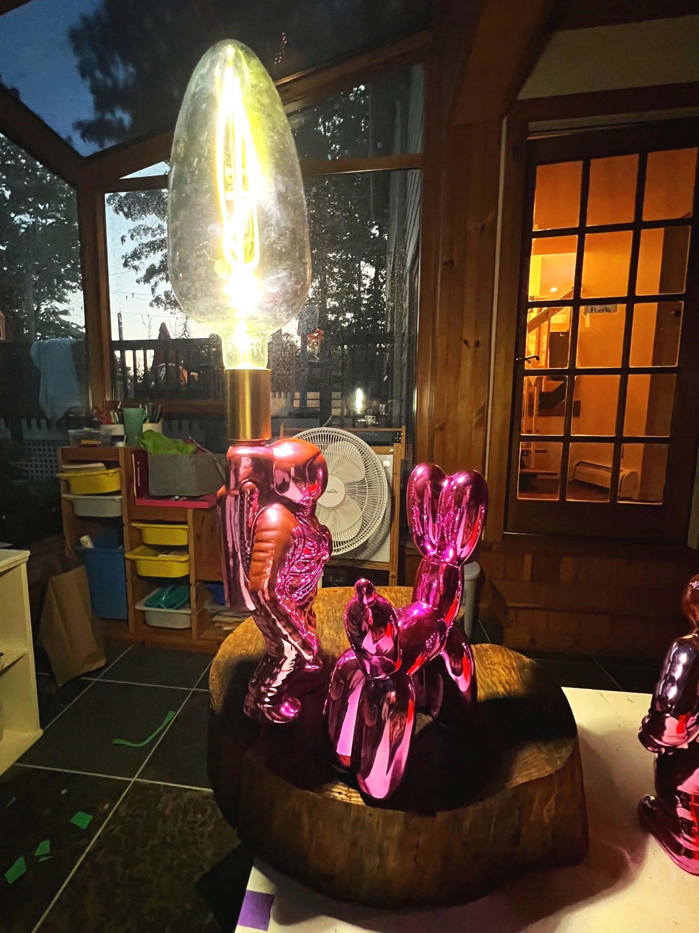 Decorative pink glass sculptures resembling rabbits and carrots on a wooden base, illuminated by a large decorative light bulb, in a cozy indoor space with wooden walls and a window showing the outdoors.