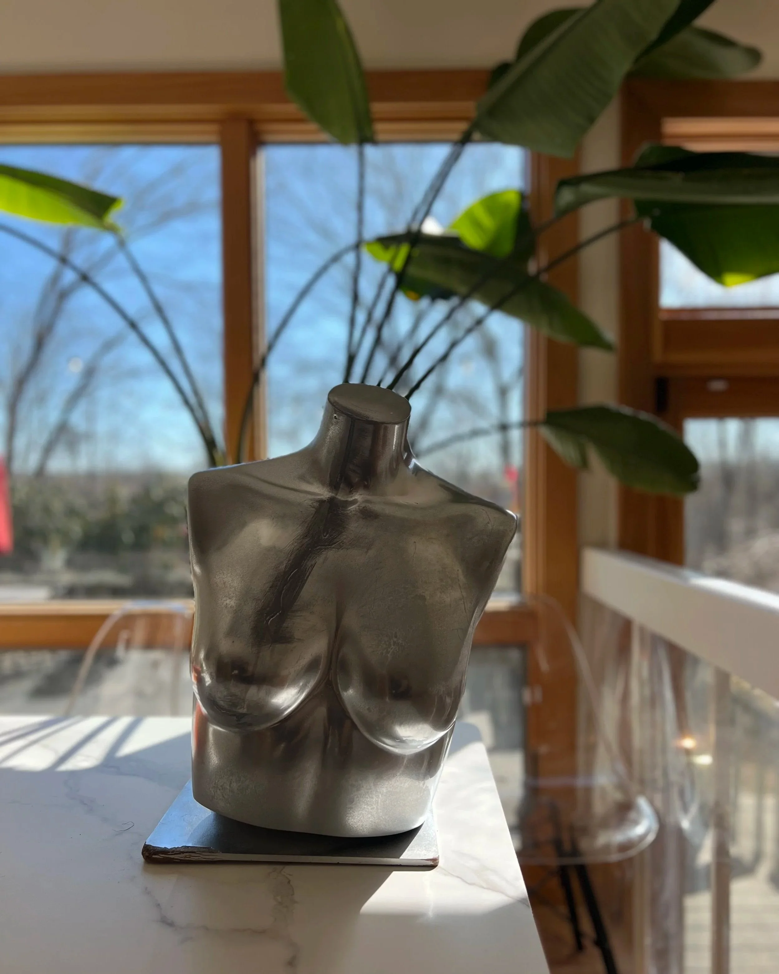 Metallic torso sculpture of a female bust placed on a white marble surface in front of a window with a large green indoor plant behind it.