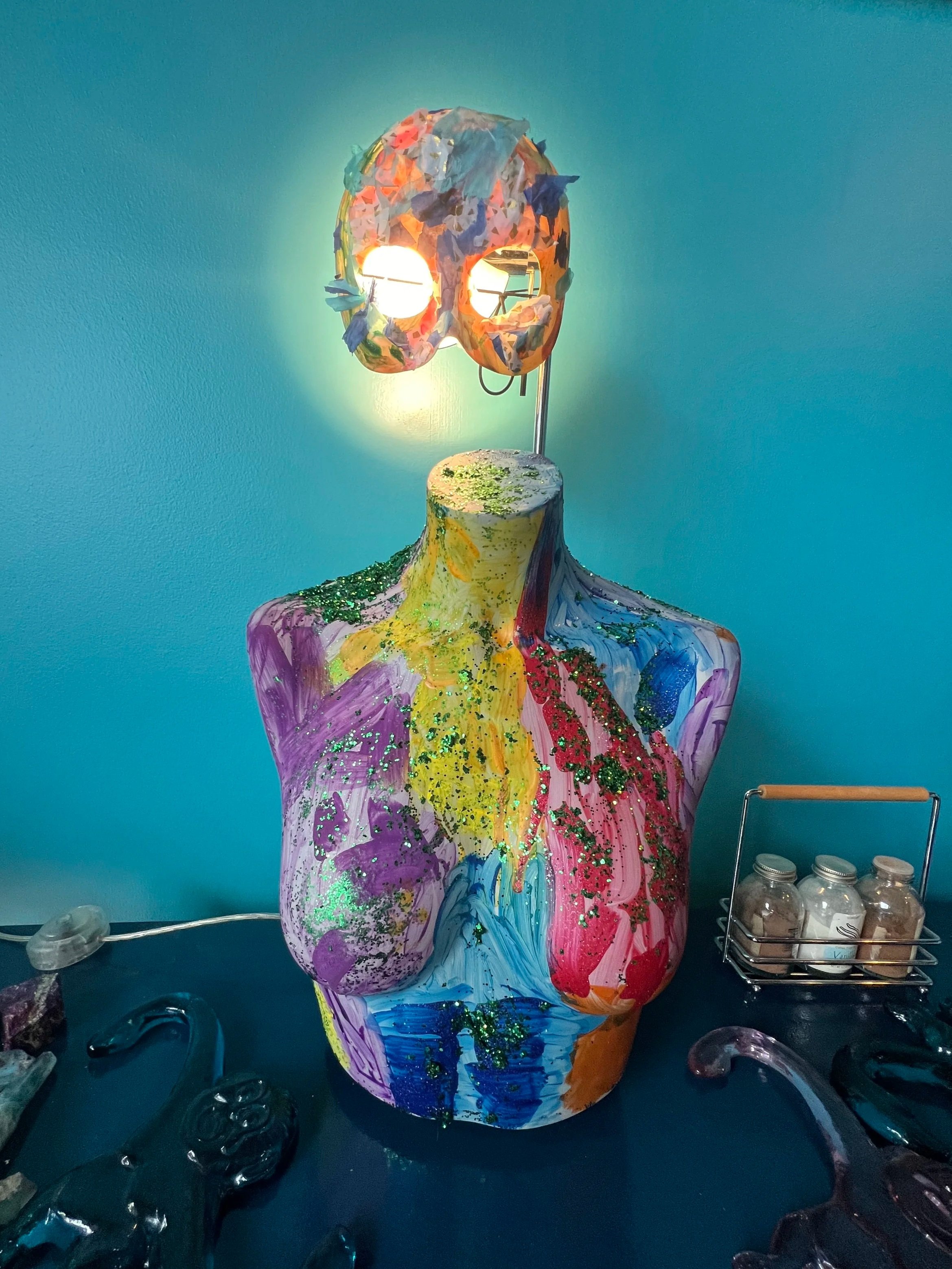 Colorful mannequin bust with painted design and glitter, topped with a paper mask illuminated from behind.