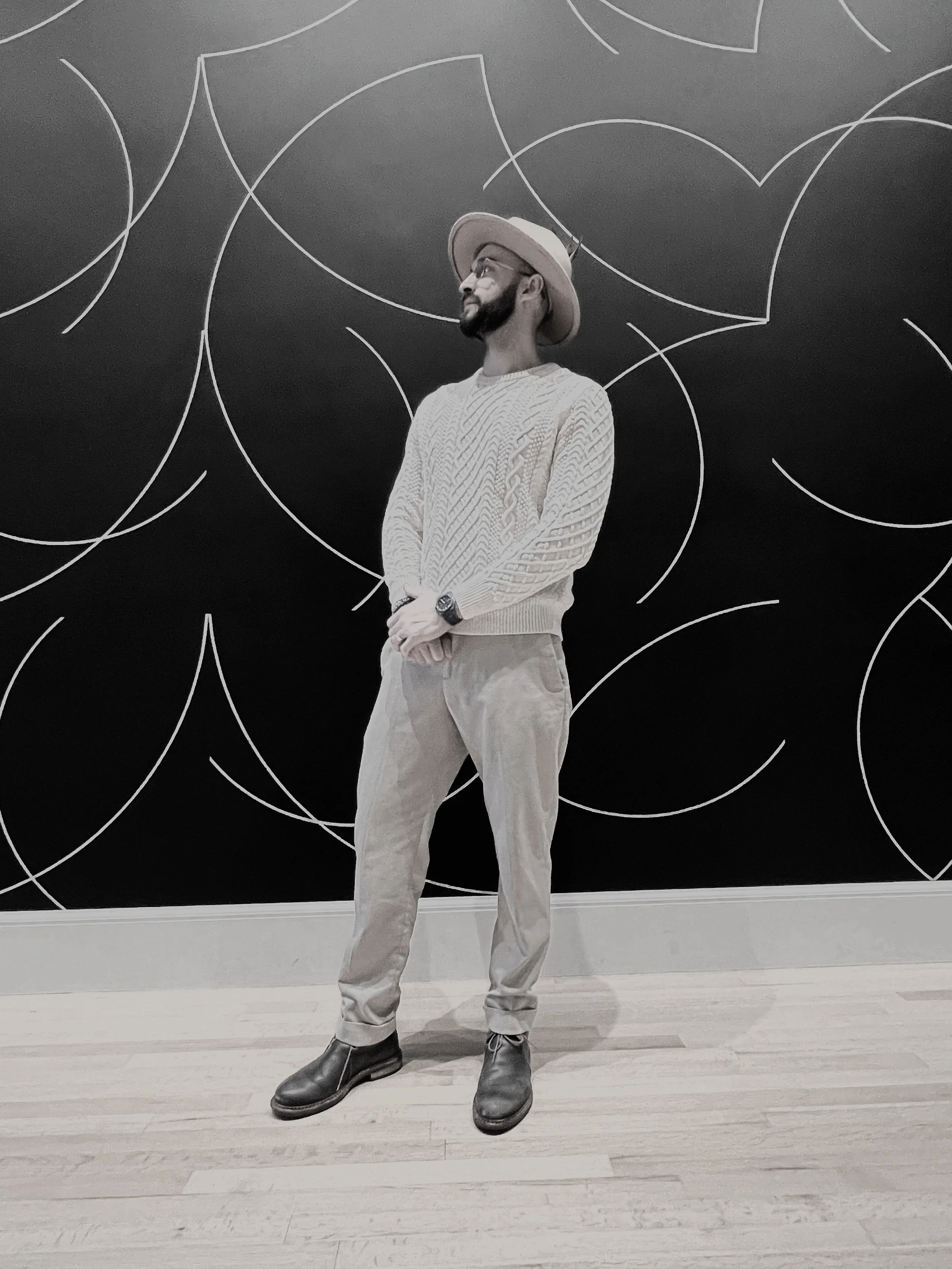 A man standing with his hands clasped in front of him, wearing a wide-brimmed hat, glasses, a light-colored cable-knit sweater, beige pants, and dark boots, posed in front of a black wall with white geometric circular patterns.