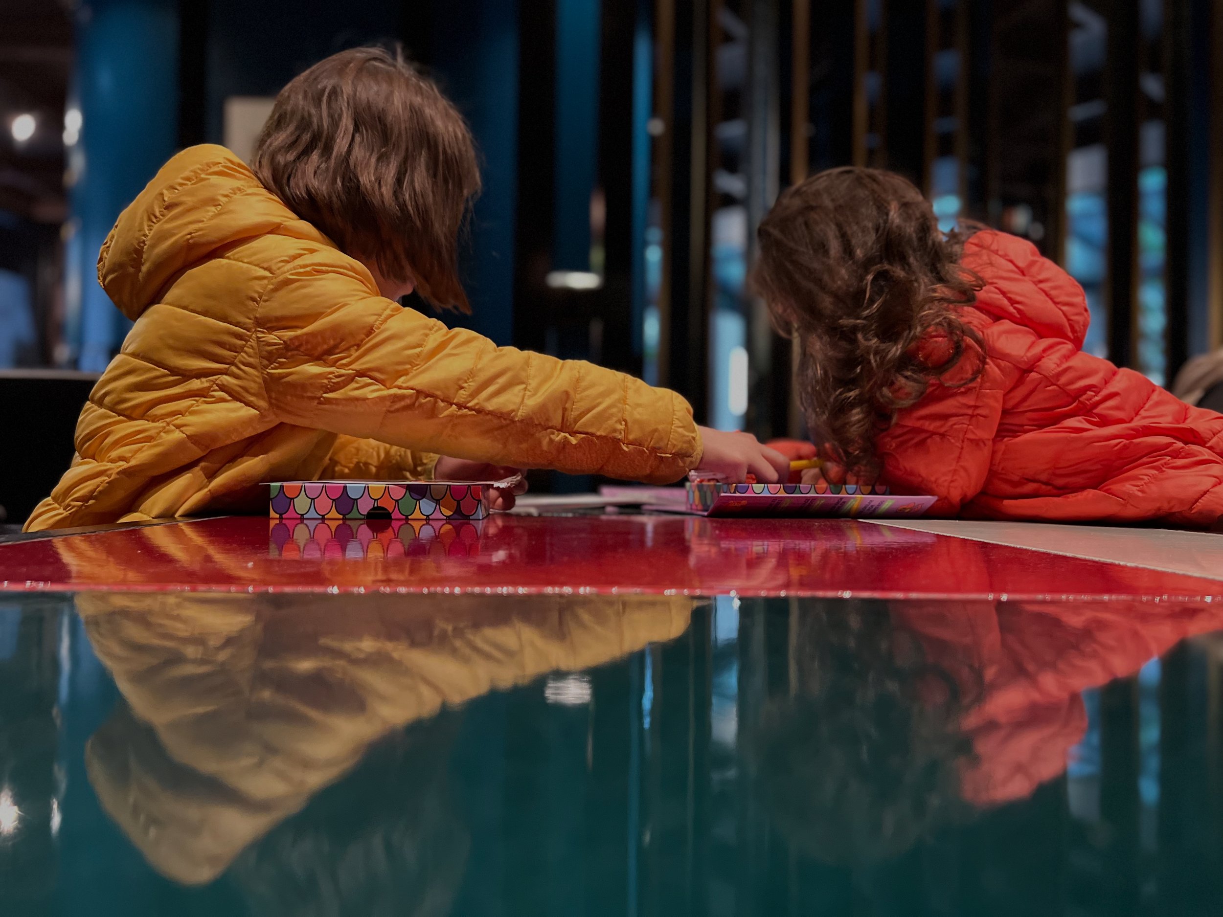 Two children in orange and yellow puffer jackets leaning over a table, reaching for objects in colorful boxes, with their reflections visible on the shiny surface of the table.